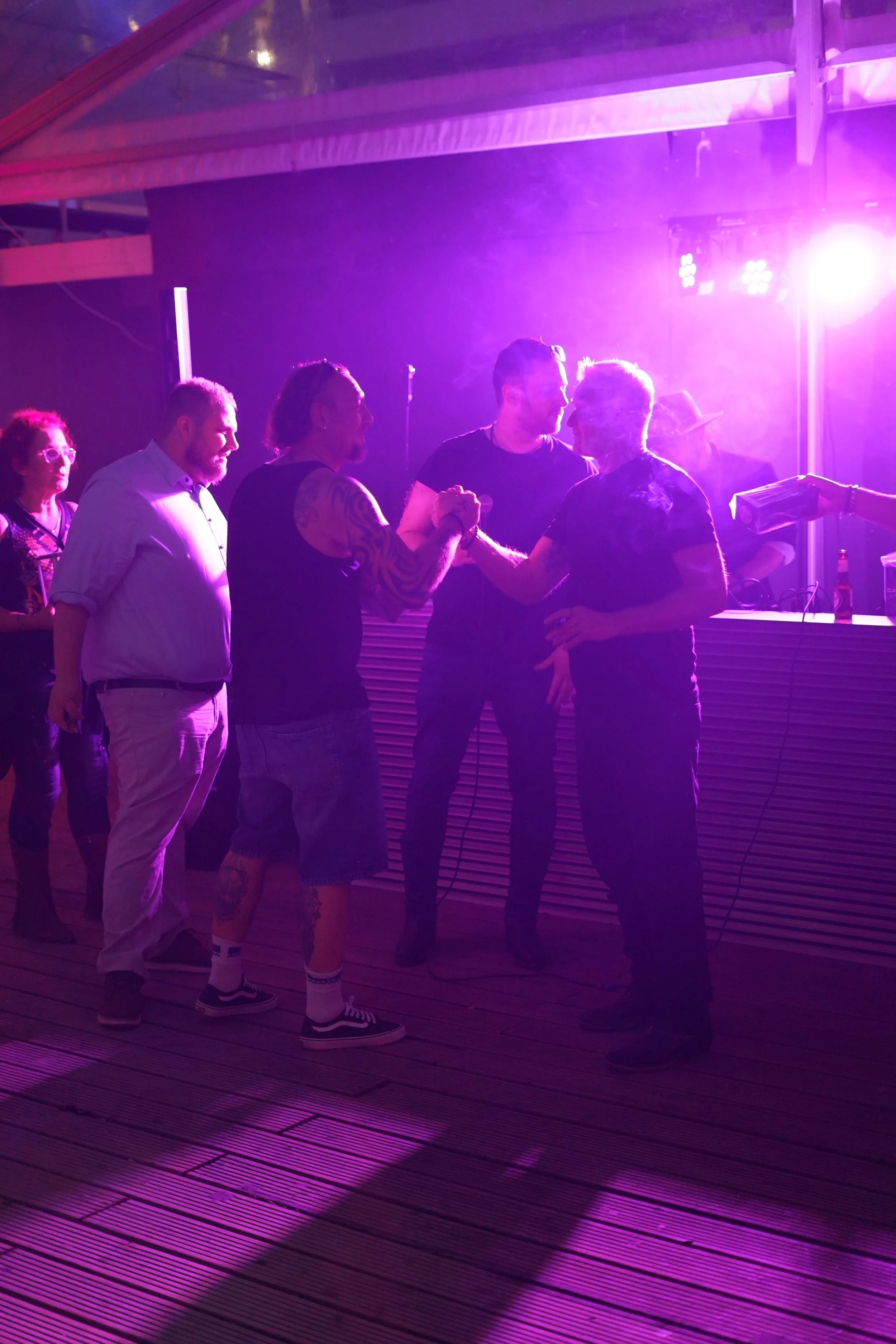 Group of people at a social gathering under purple and pink lighting, two men are shaking hands and others are standing nearby.