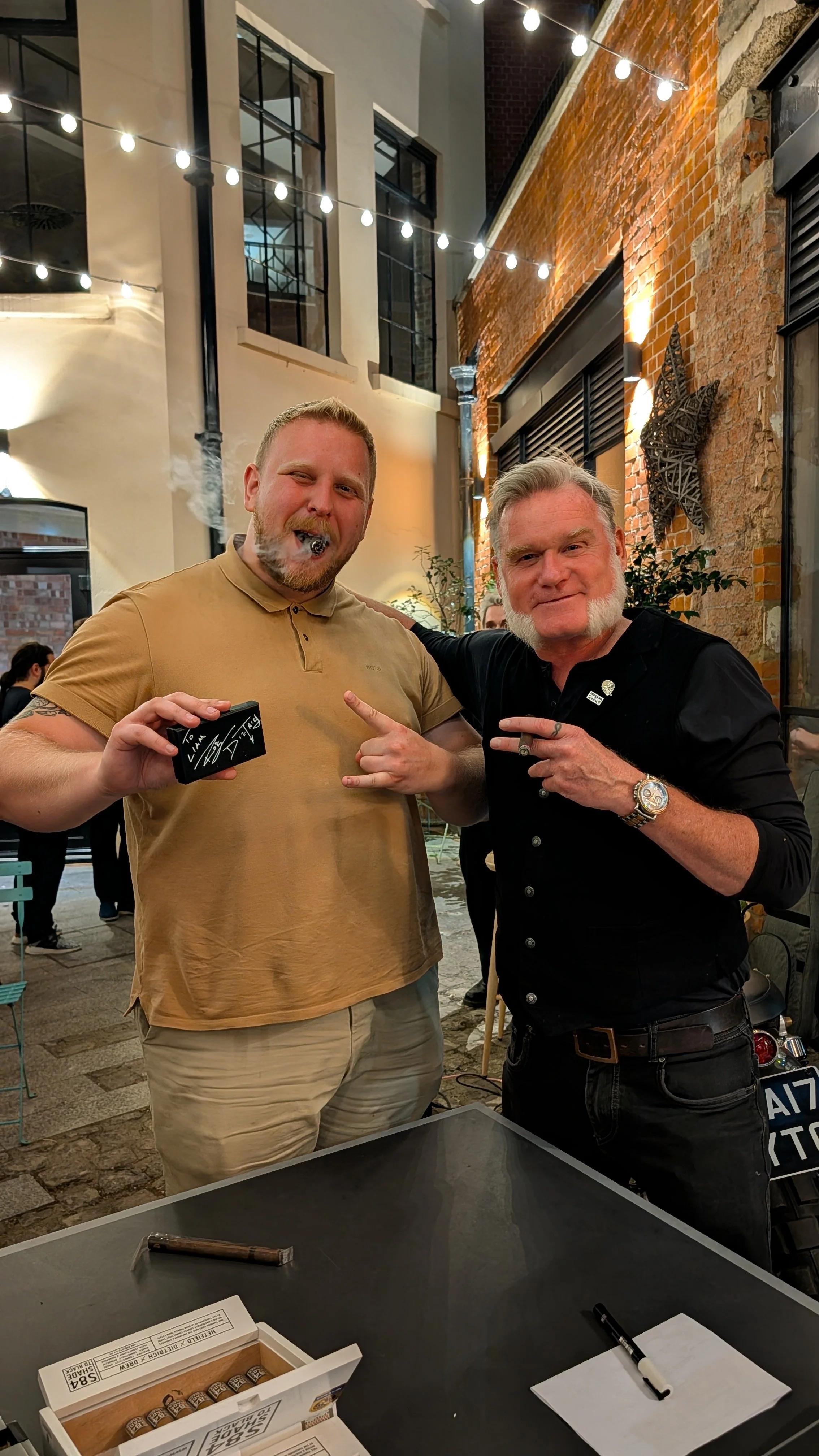 Two men standing outside at night, smiling and posing with one making a rock sign, with string lights above and a brick wall behind them.