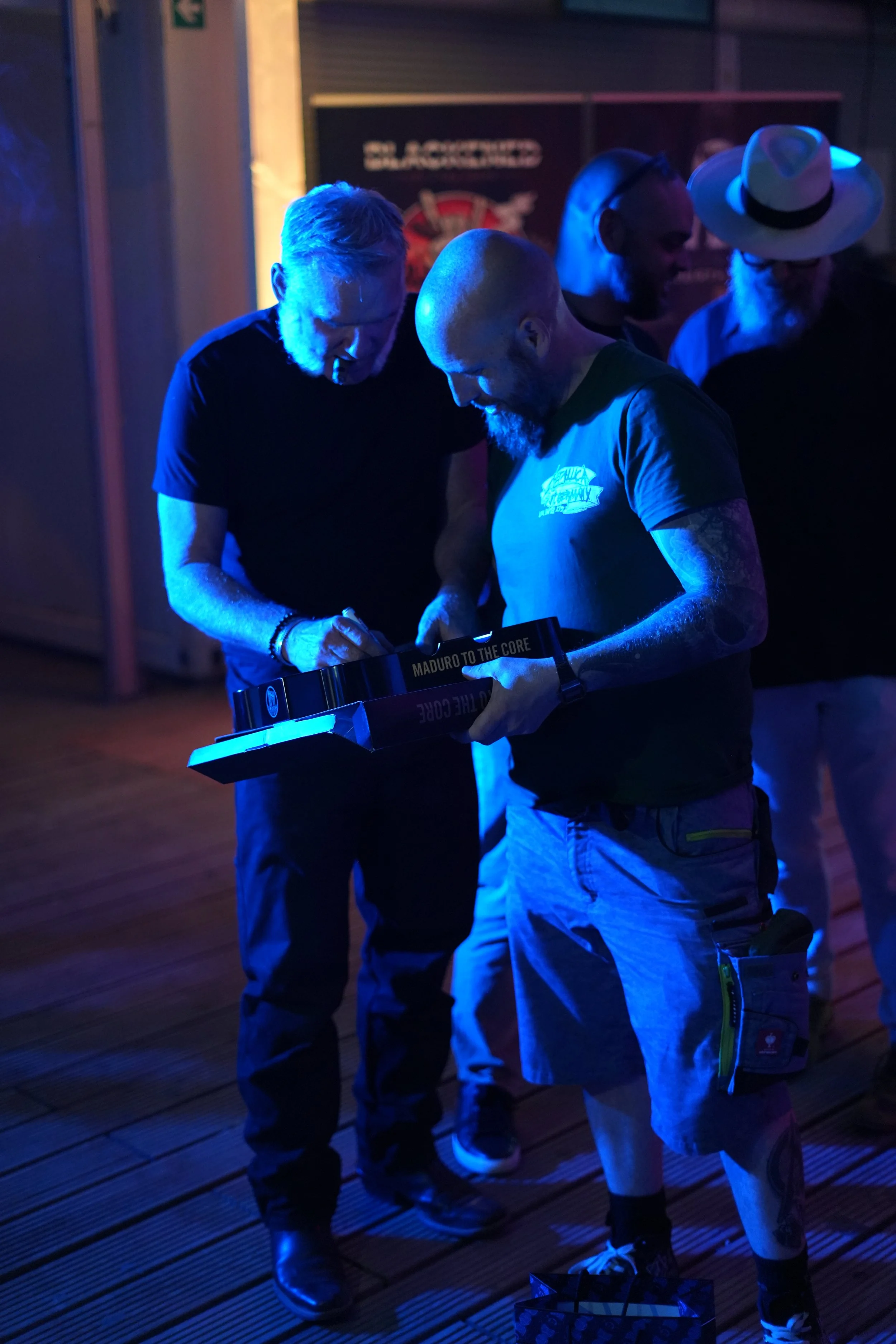 Three men are gathered around a small electronic or DJ equipment in a dimly lit environment with blue and purple lighting. One man is wearing a light-colored t-shirt with tattoos on his arms, another is wearing a dark shirt, and the third is wearing 