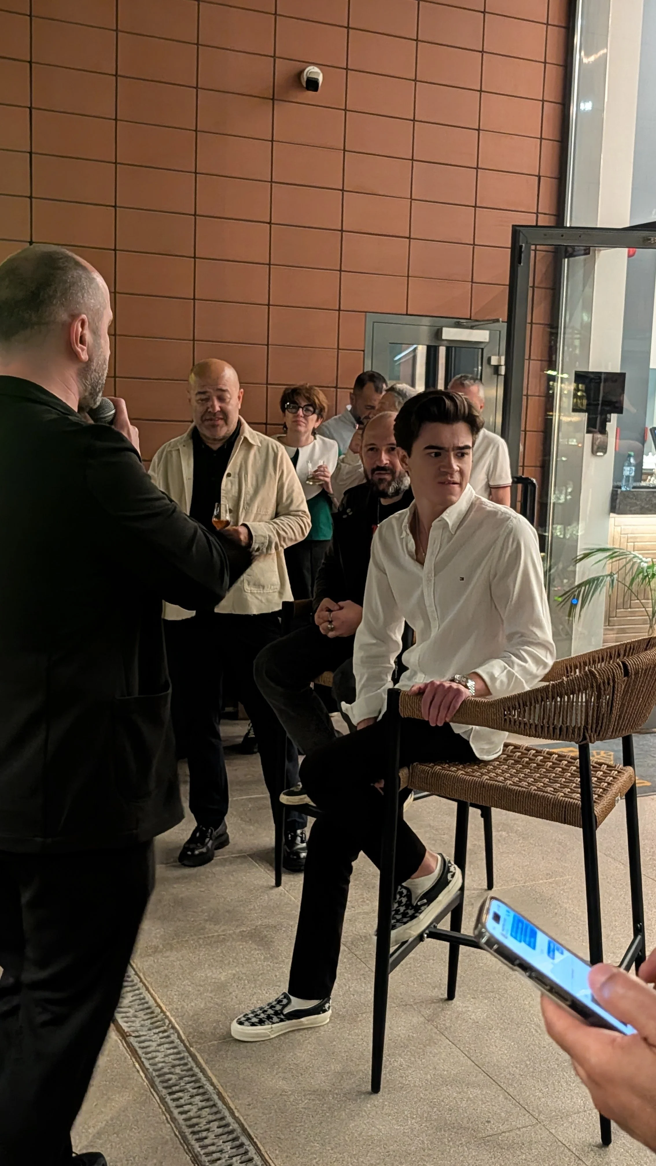 A man with a phone showing a barcode is sitting on a high stool at what appears to be a social gathering or event. Other people are standing and sitting nearby, some holding drinks, inside a modern indoor space with brick walls and large glass doors.