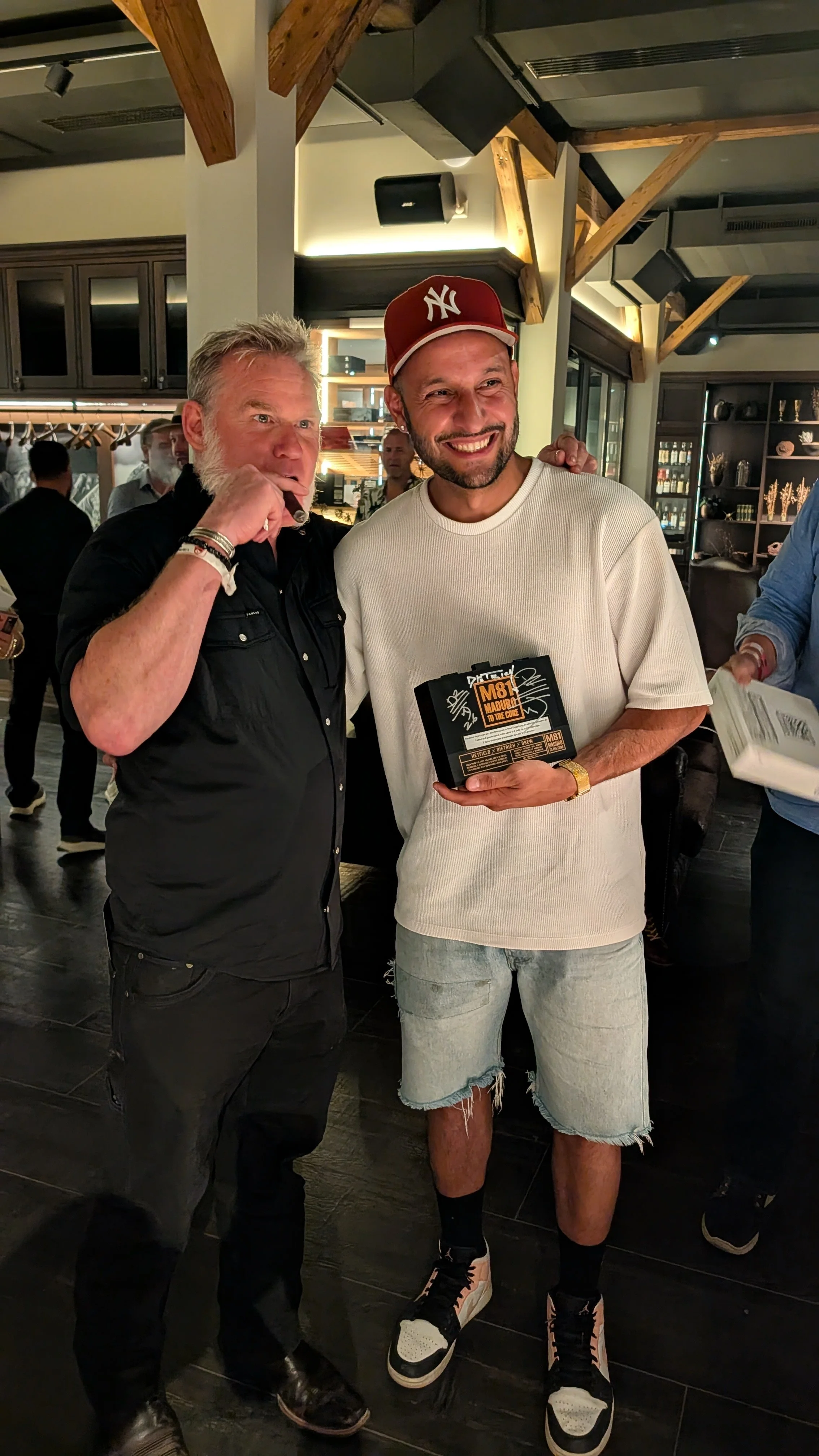 Two men are inside a restaurant, one holding what appears to be a box or award, and the other making a speech or gesture. The man on the right is wearing a white T-shirt, denim shorts, a red New York Yankees cap, and sneakers. The man on the left is 