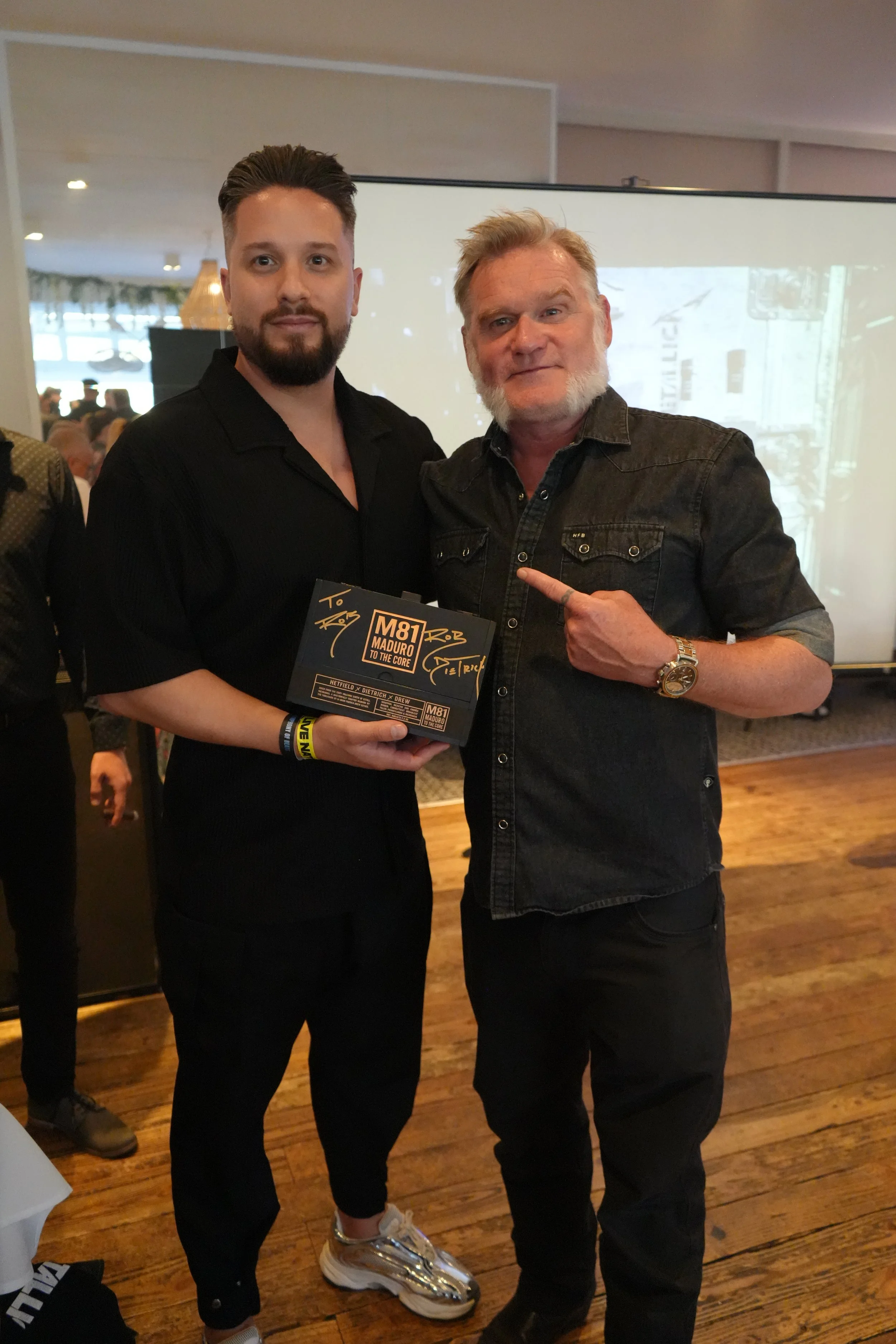 Two men standing indoors, one holding a signed poster. The man on the left has dark hair and beard, wearing a black shirt and sneakers. The man on the right has light hair and beard, wearing a black denim jacket and a gold watch, pointing at the post