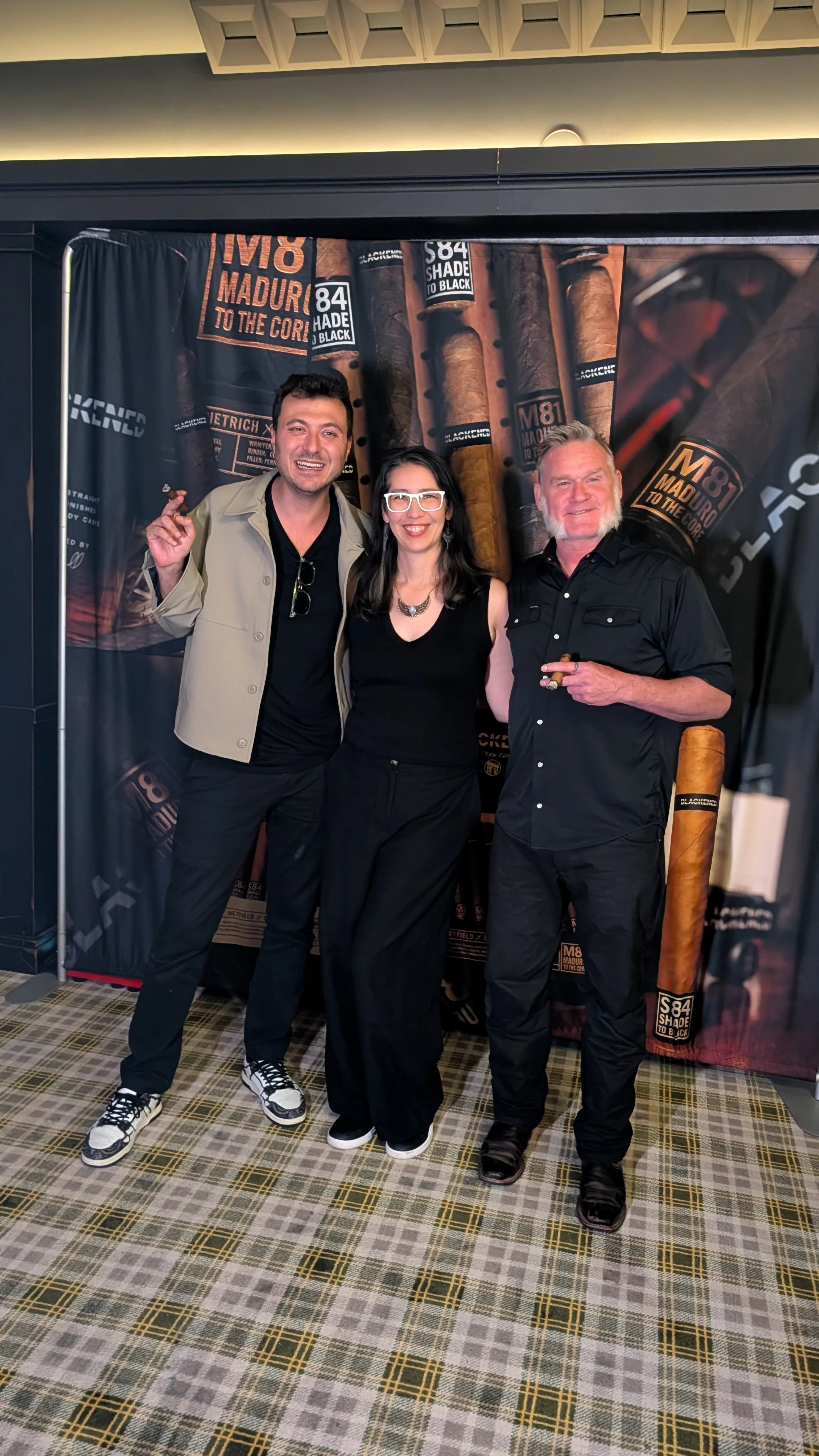 Three people standing and smiling together at an indoor event with a backdrop featuring cigars.