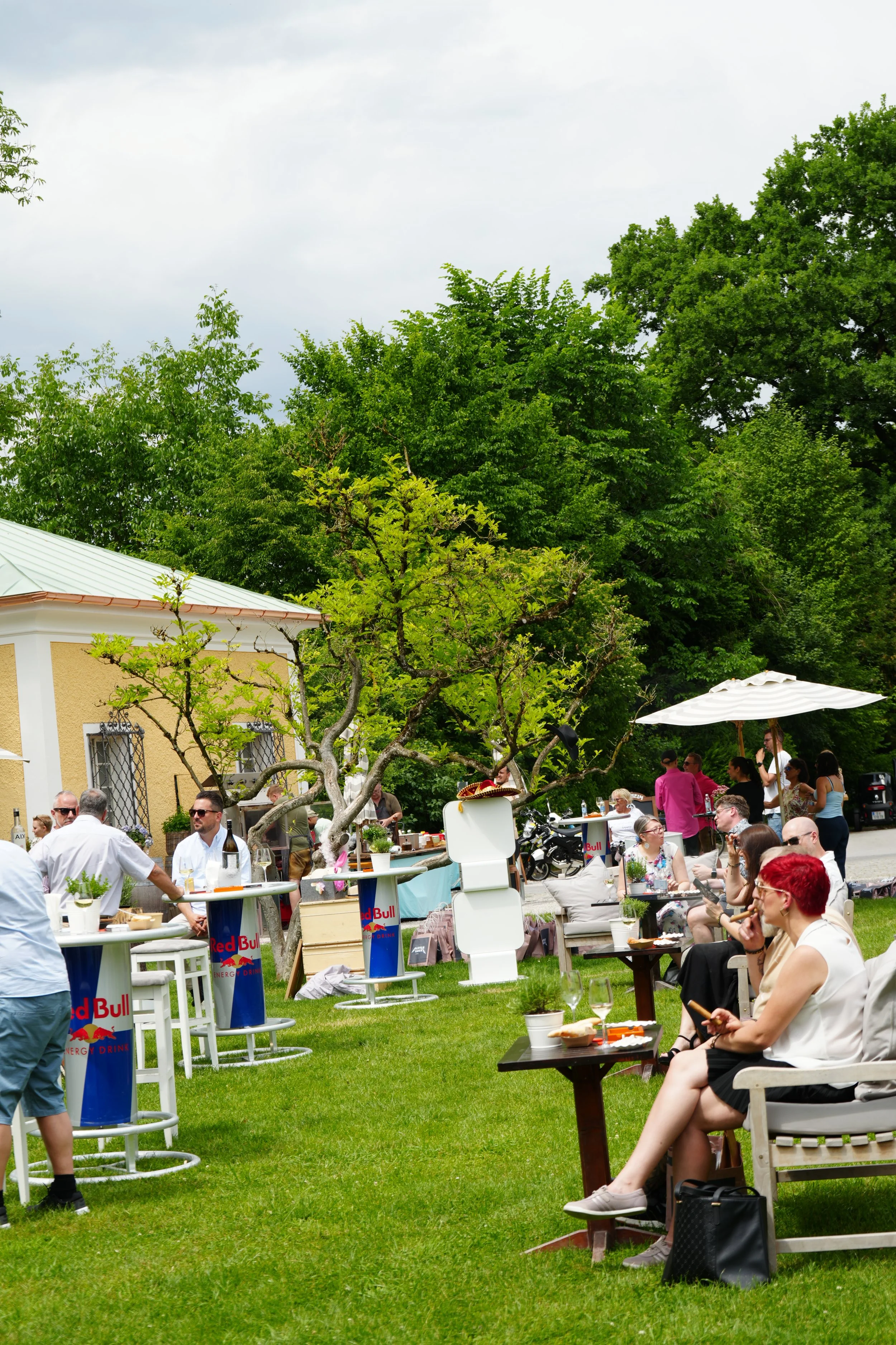 People socializing at an outdoor garden party with tables, chairs, and umbrellas, surrounded by trees and greenery on a cloudy day.