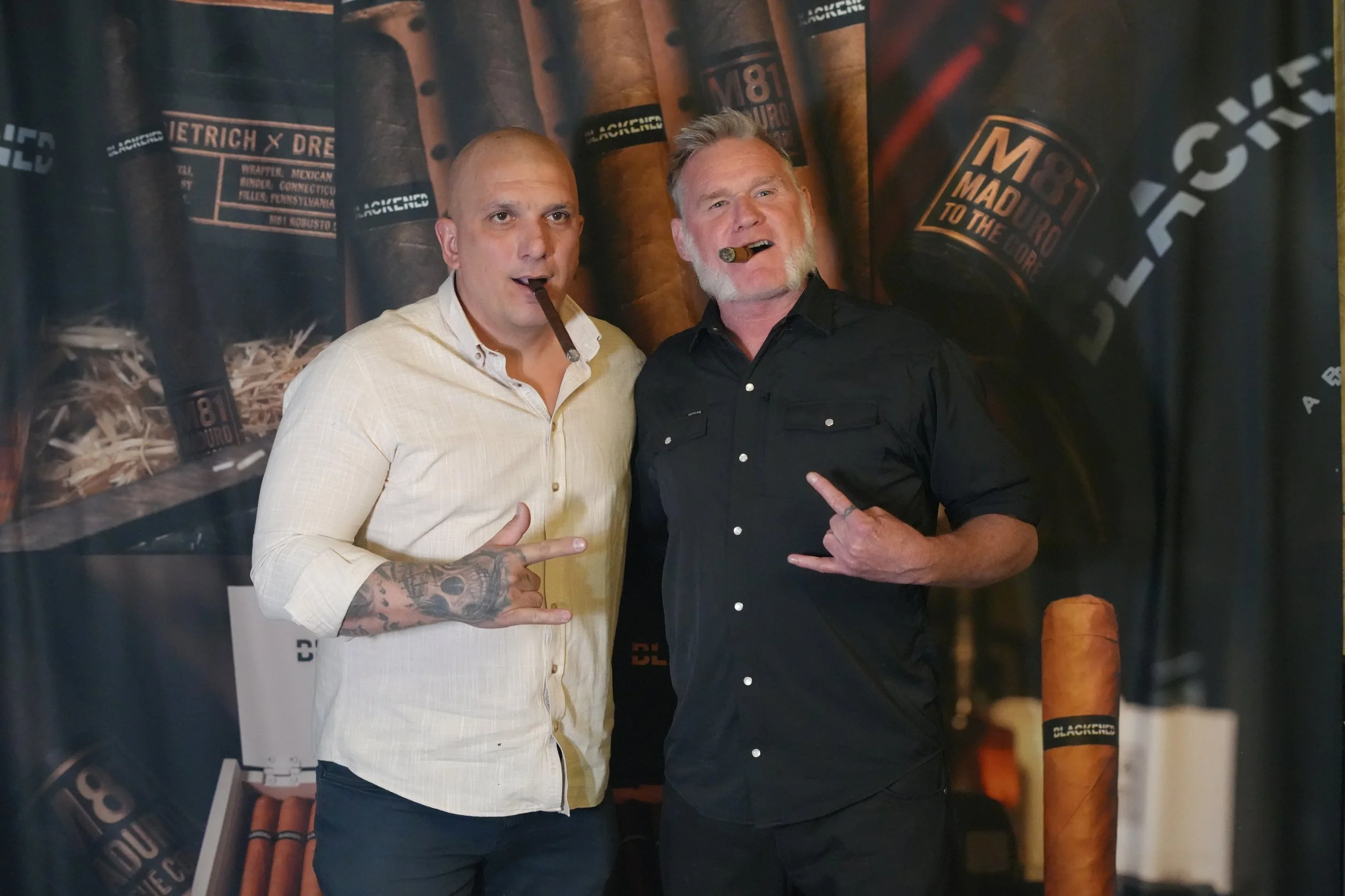Two men are posing together in front of a backdrop with large cigar images and branding. Both men have cigars in their mouths, making a shaka sign with their hands. One man has a shaved head and tattoos on his arm, wearing a cream-colored shirt. The 