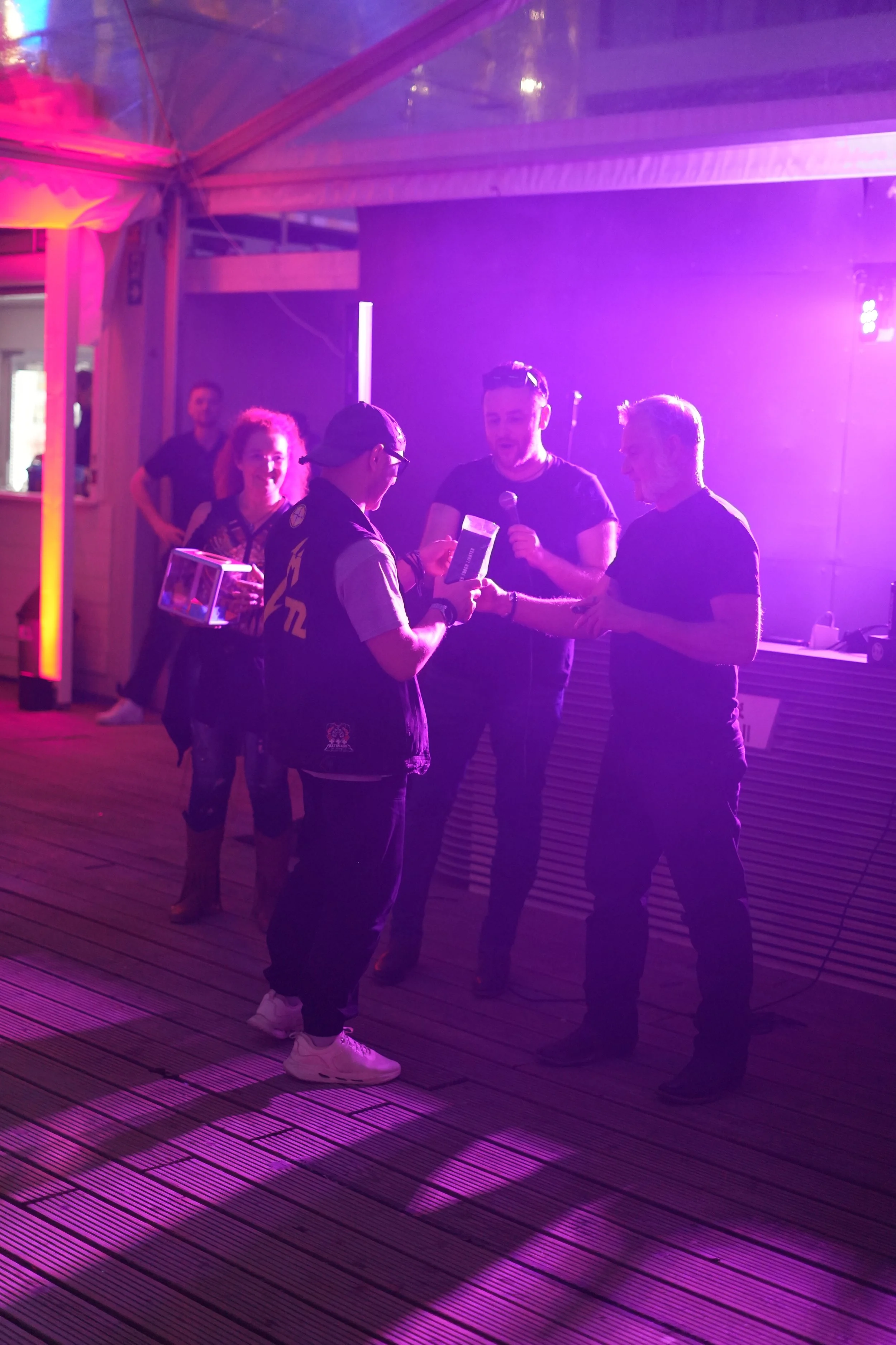 Group of people at a party or event with purple lighting, some holding drinks, engaging in conversation.