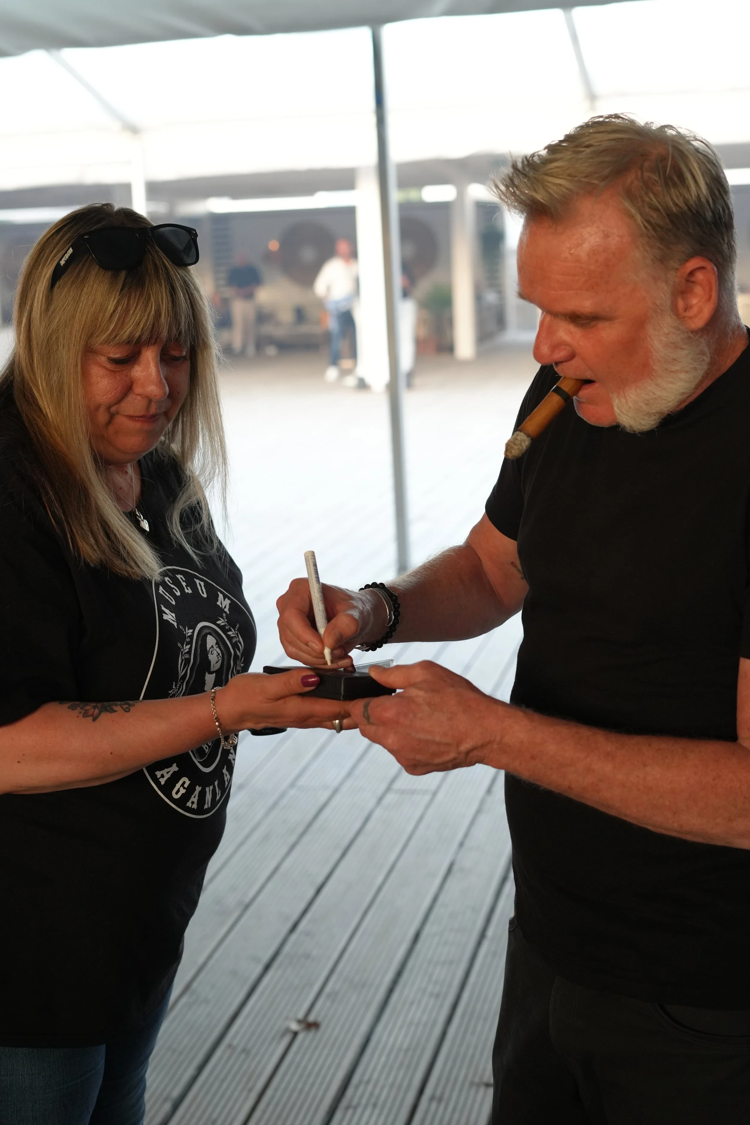 A man with a cigar in his mouth signs an object while a woman watches and holds it.