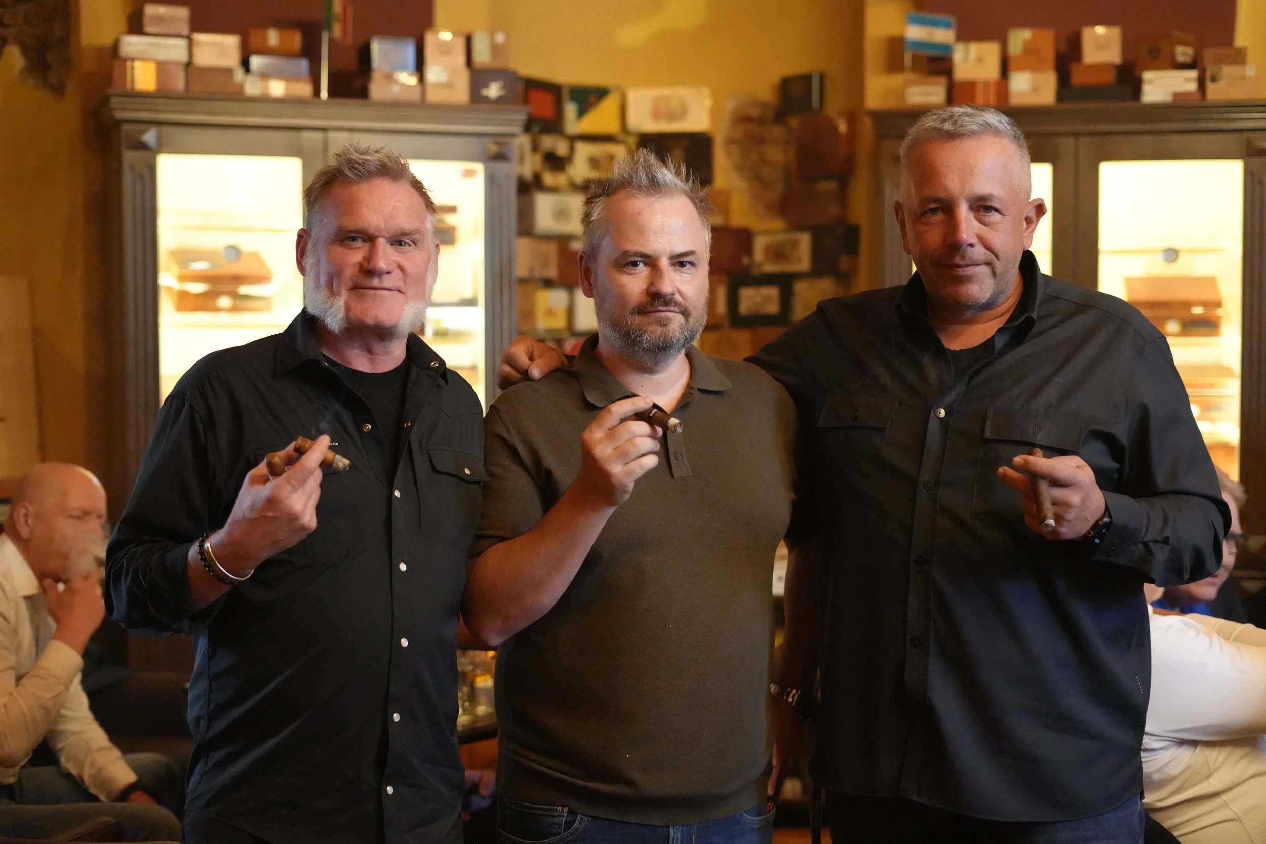 Three men standing together at a social gathering, each holding a cigar, in a warmly lit room with shelves of boxes and other people in the background.