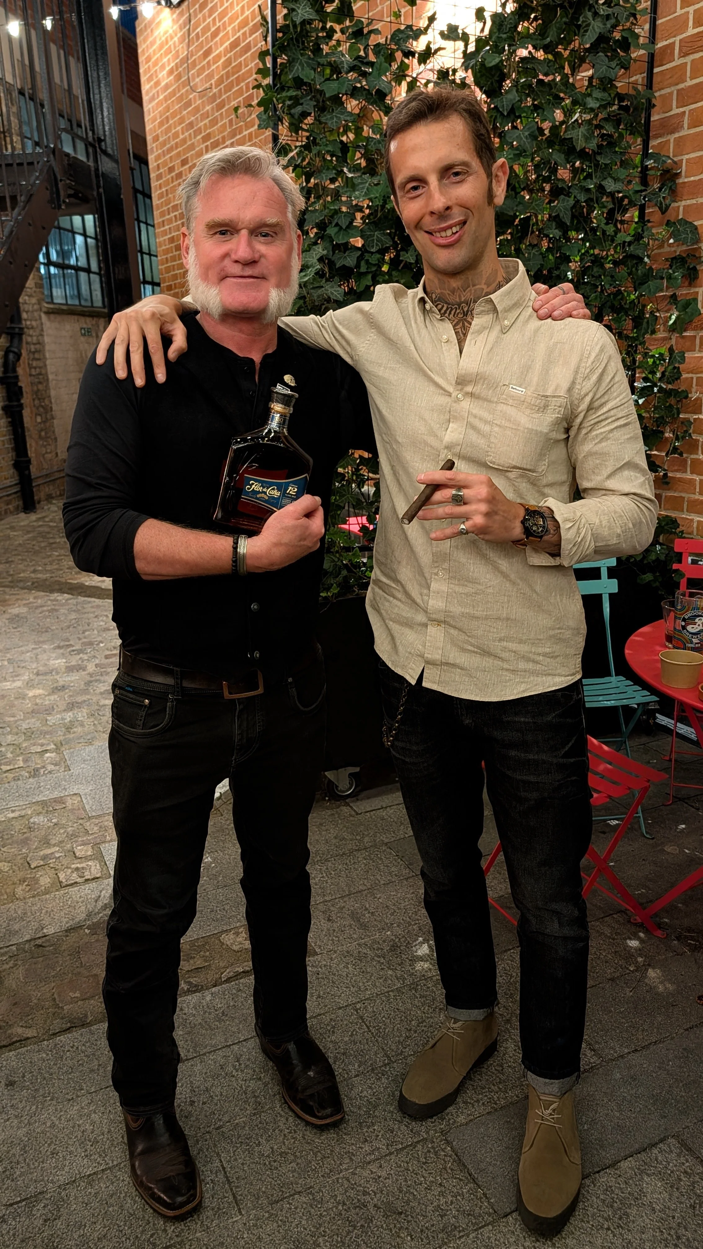 Two men standing outside near brick walls, one holding a bottle of alcohol and the other holding a cigar. Both smiling, with one placing a hand on the other's shoulder.