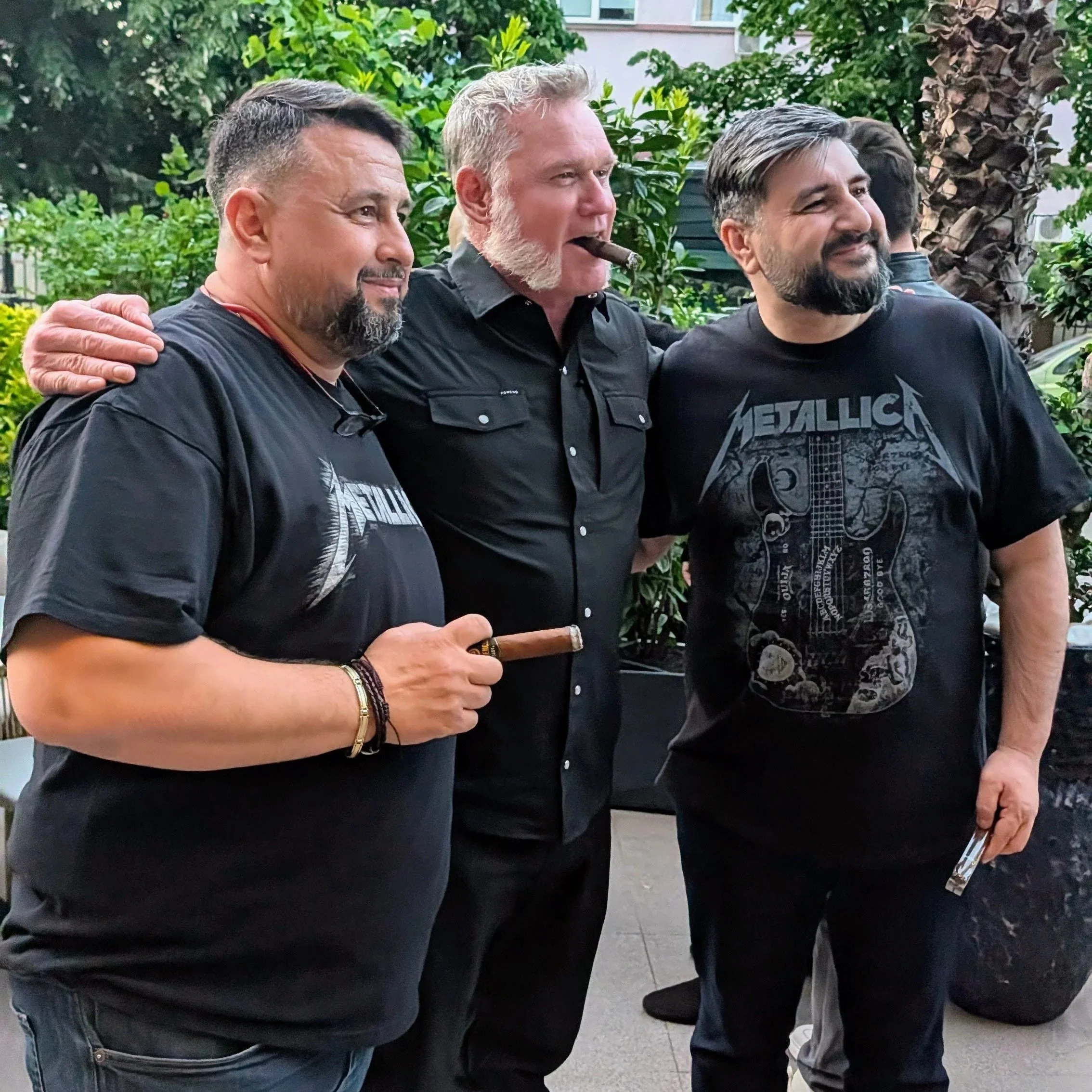 Three men standing outdoors in front of green foliage. The man in the middle has white hair and a beard, is wearing a black shirt, and is smoking a cigar. The men on either side have darker hair and beards, wearing black shirts, and are smiling. One of them is wearing a Metallica t-shirt.