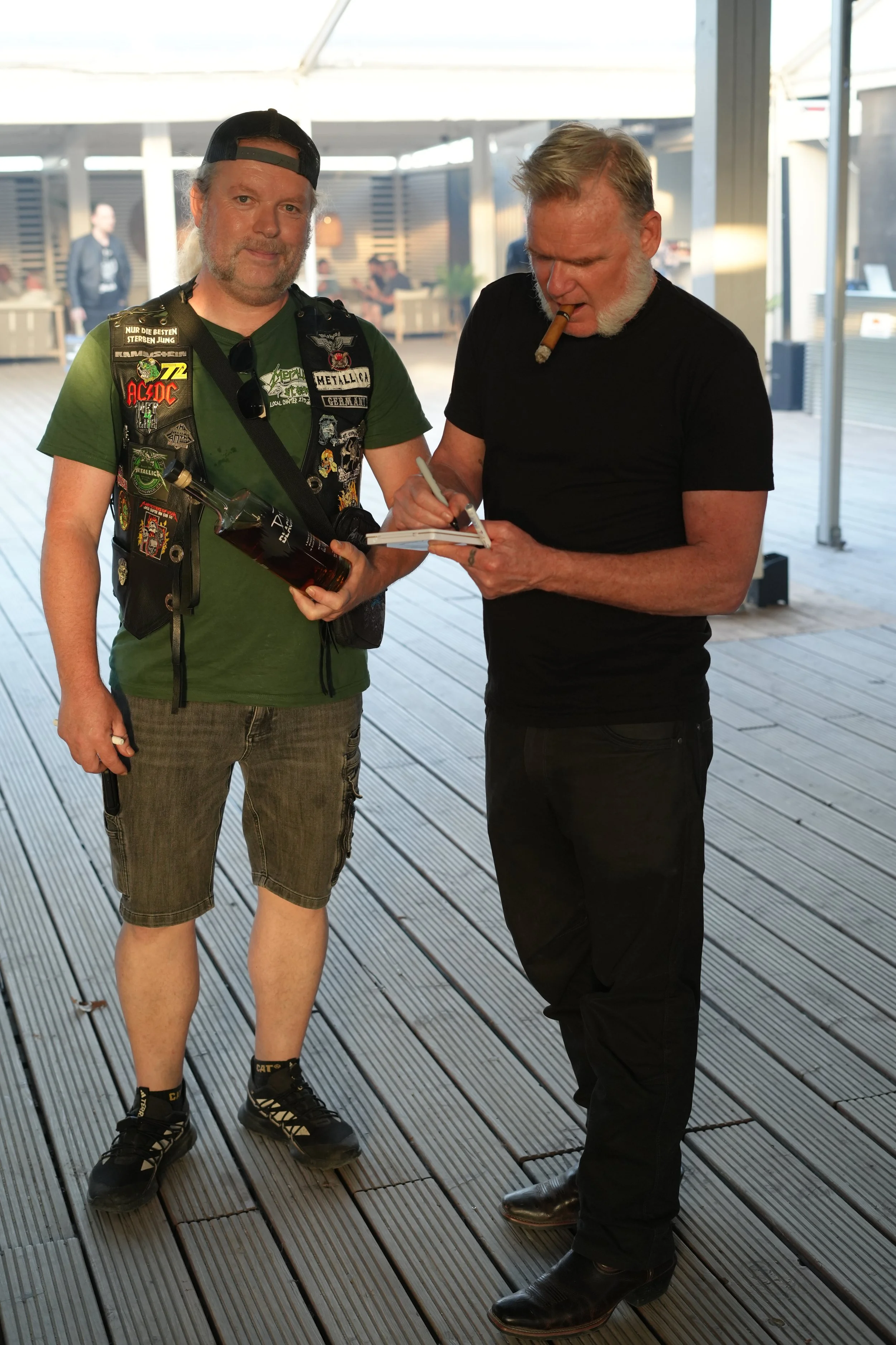 Two men standing on a wooden deck area, one holding a bottle of alcohol and the other signing an autograph on a notepad. The man on the right has a cigar in his mouth and is dressed in black, while the man on the left is wearing casual attire and a v
