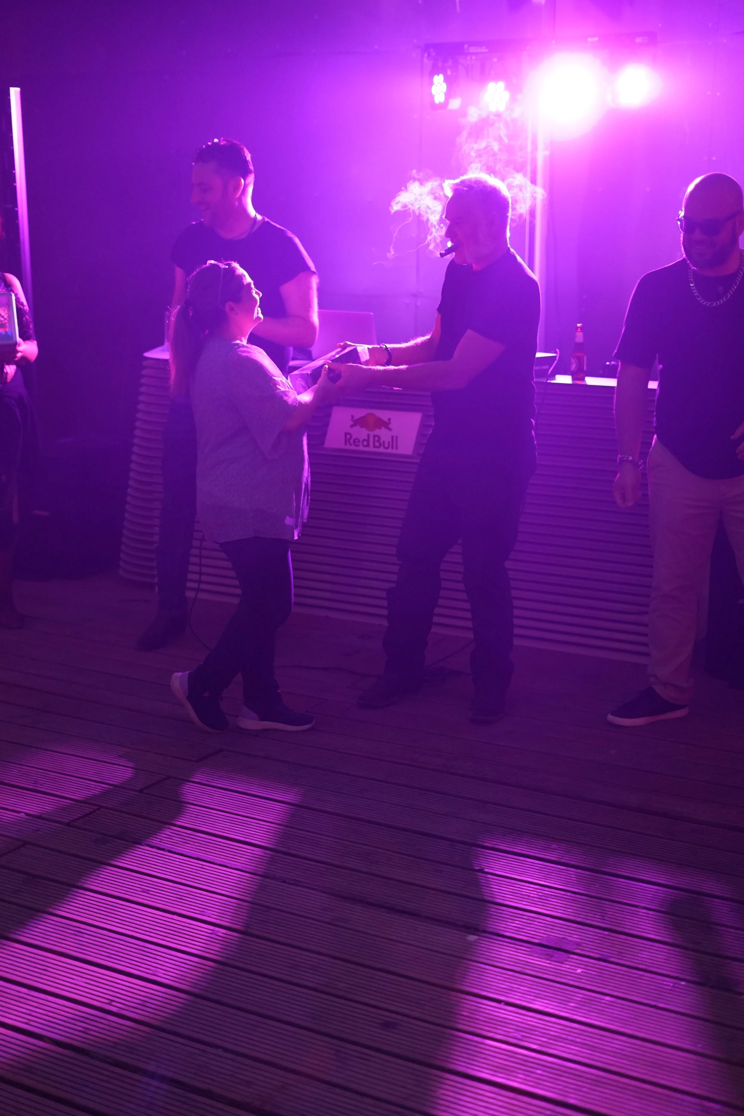 People at a nightclub under purple and pink lighting, with a DJ or performer behind a Red Bull bar, engaging with a young girl.