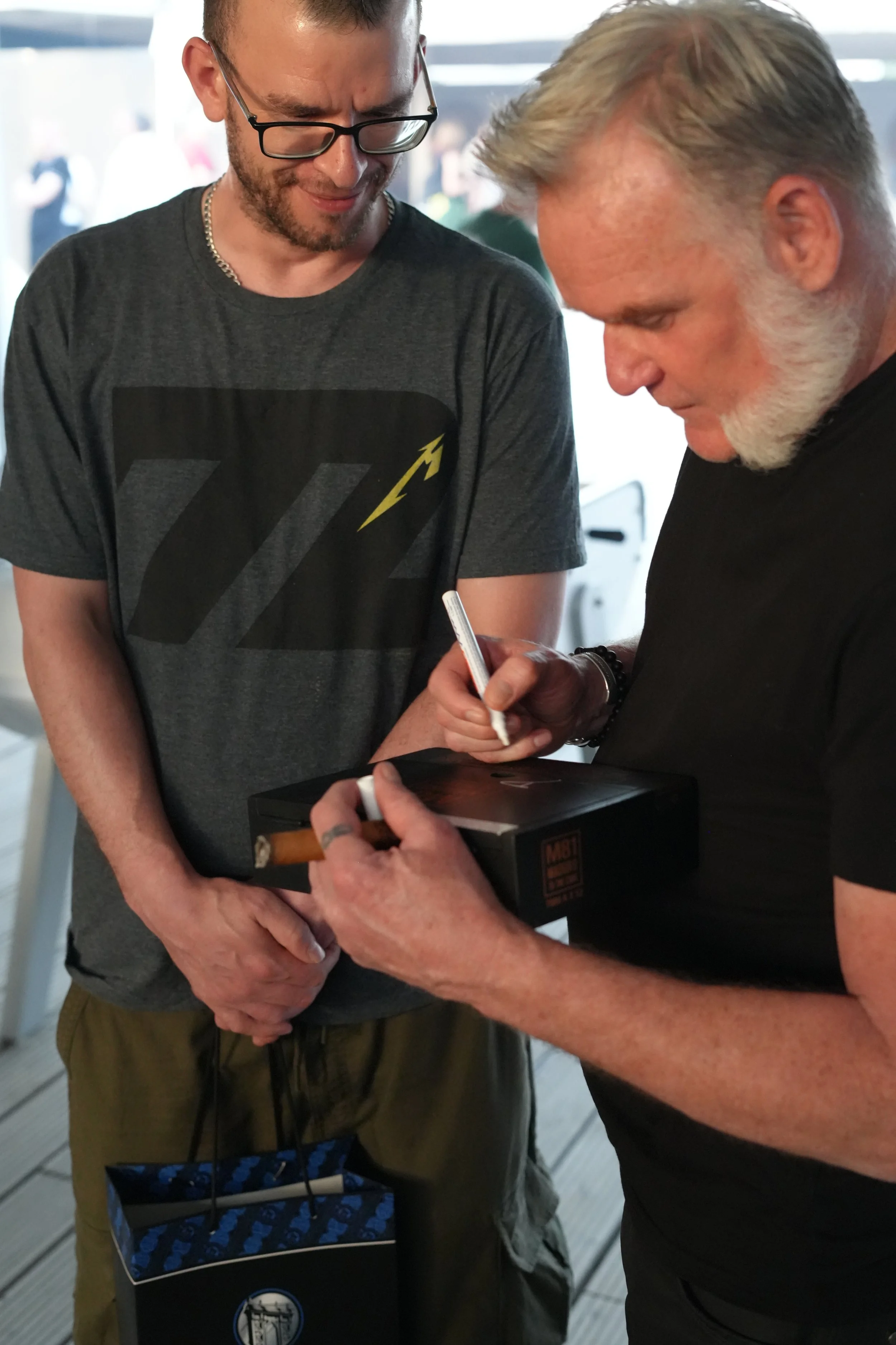 Two men are engaged in a conversation; one is signing an item while holding a cigar, the other is looking down at the signing. One man has glasses, a beard, and short hair, and the other has gray hair and a beard. They are standing indoors with a win