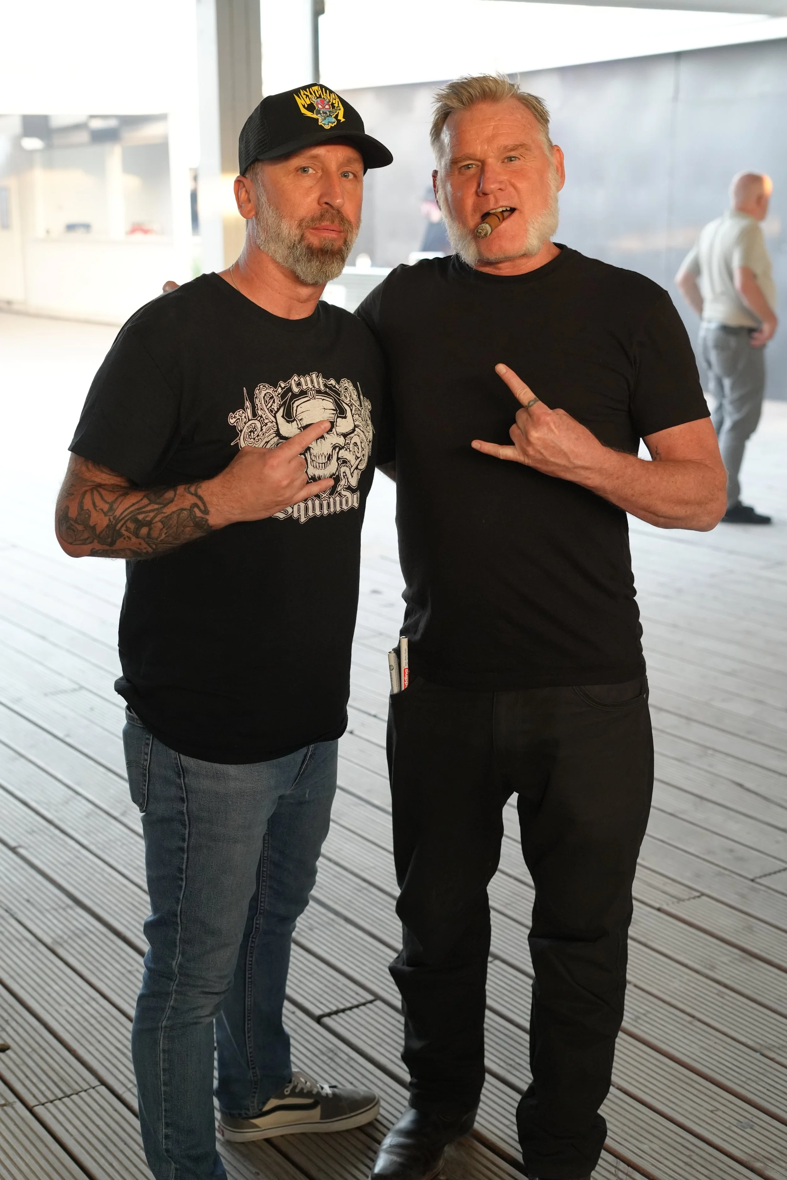 Two men standing together on a wooden deck, one wearing a black t-shirt with a skull design and a black cap, the other with a cigar in his mouth making a rock sign with his hand, both wearing black t-shirts.