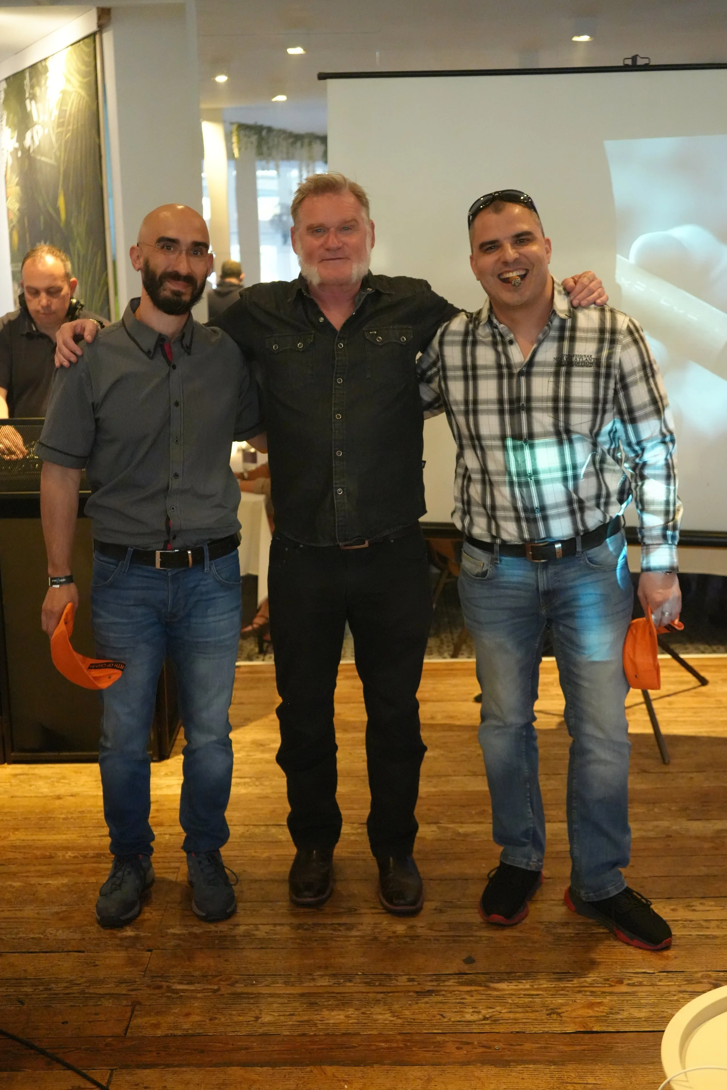 Three men standing together indoors, smiling, with a projector screen behind them and wooden flooring.