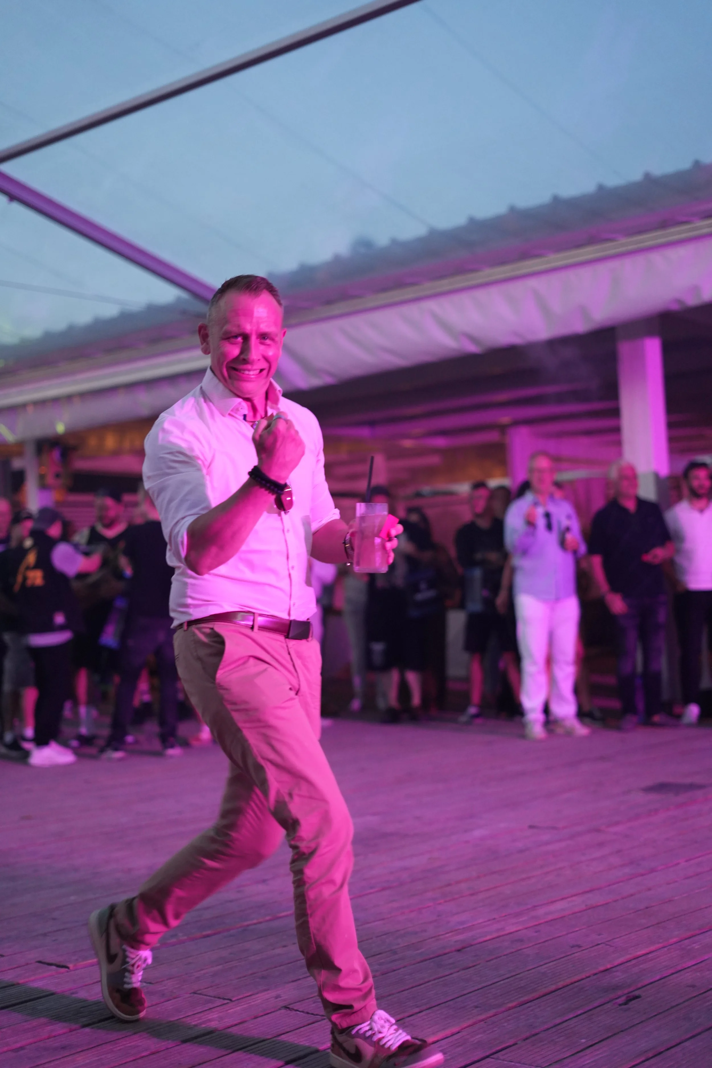 A man in a light pink shirt and beige pants dancing or celebrating at an event with pink lighting, holding a drink and making a fist gesture, with a group of people in the background under an open ceiling.