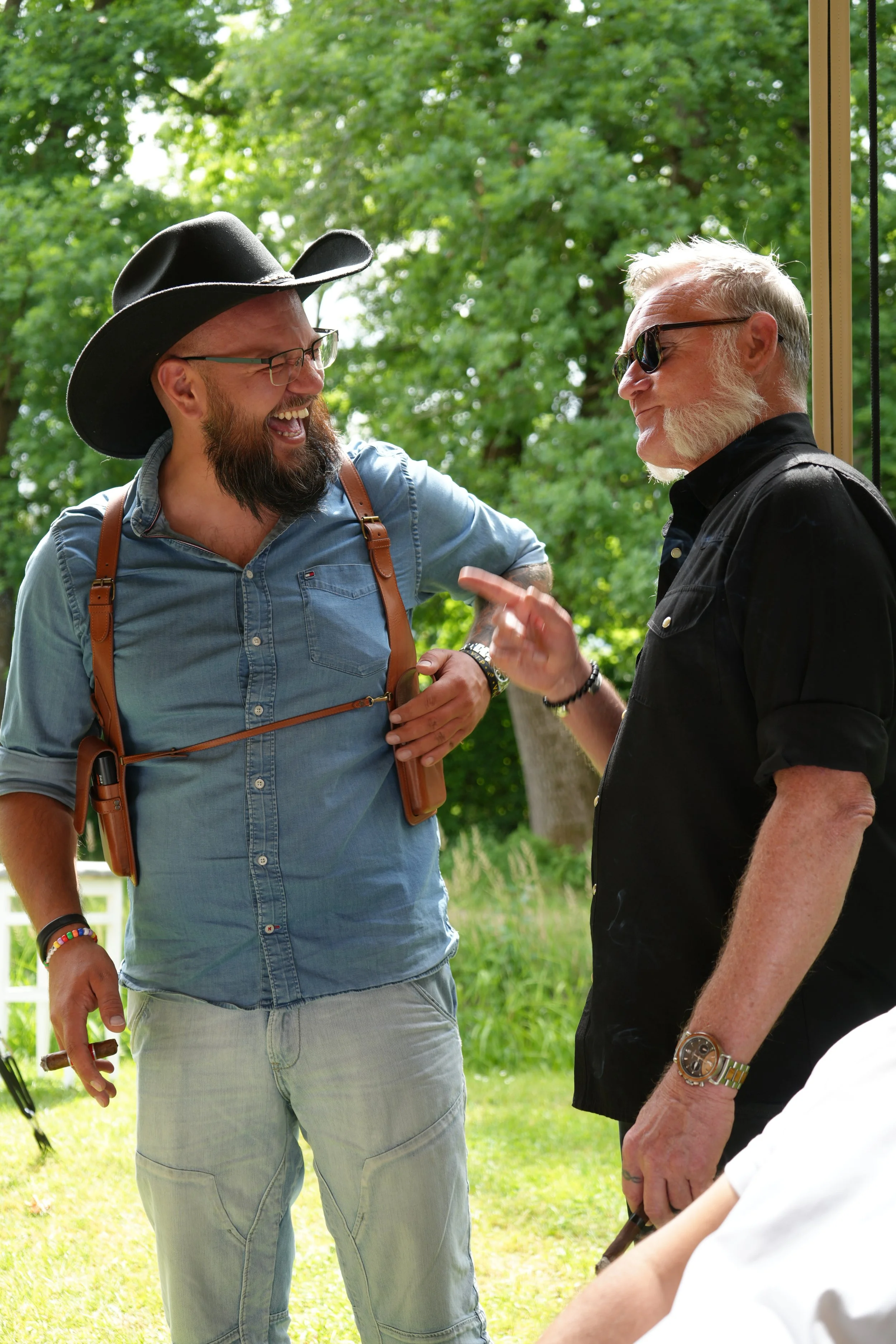 Two men having a conversation outdoors surrounded by green trees. One man is wearing a large black cowboy hat, glasses, a denim shirt, and light-colored jeans. The other man is wearing sunglasses, a black shirt, and has a white beard. They appear to 