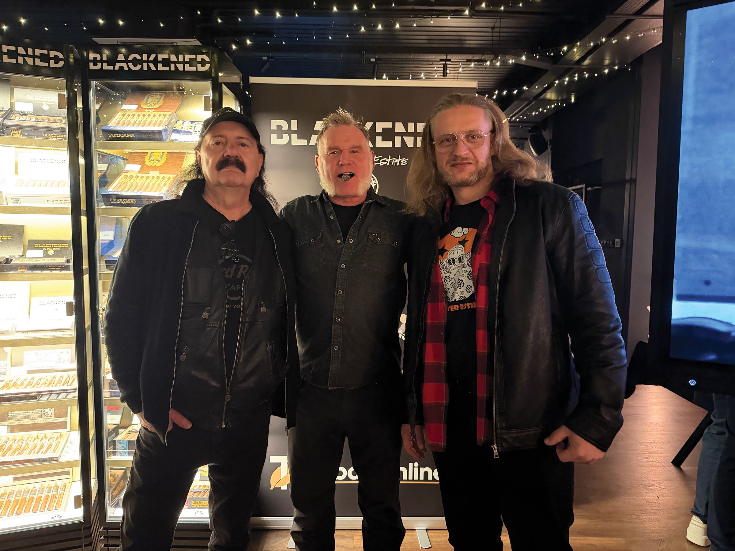 Three men standing together inside a cigar shop or lounge, with a display case of cigars and a large sign that reads 'BLACKENED' behind them.