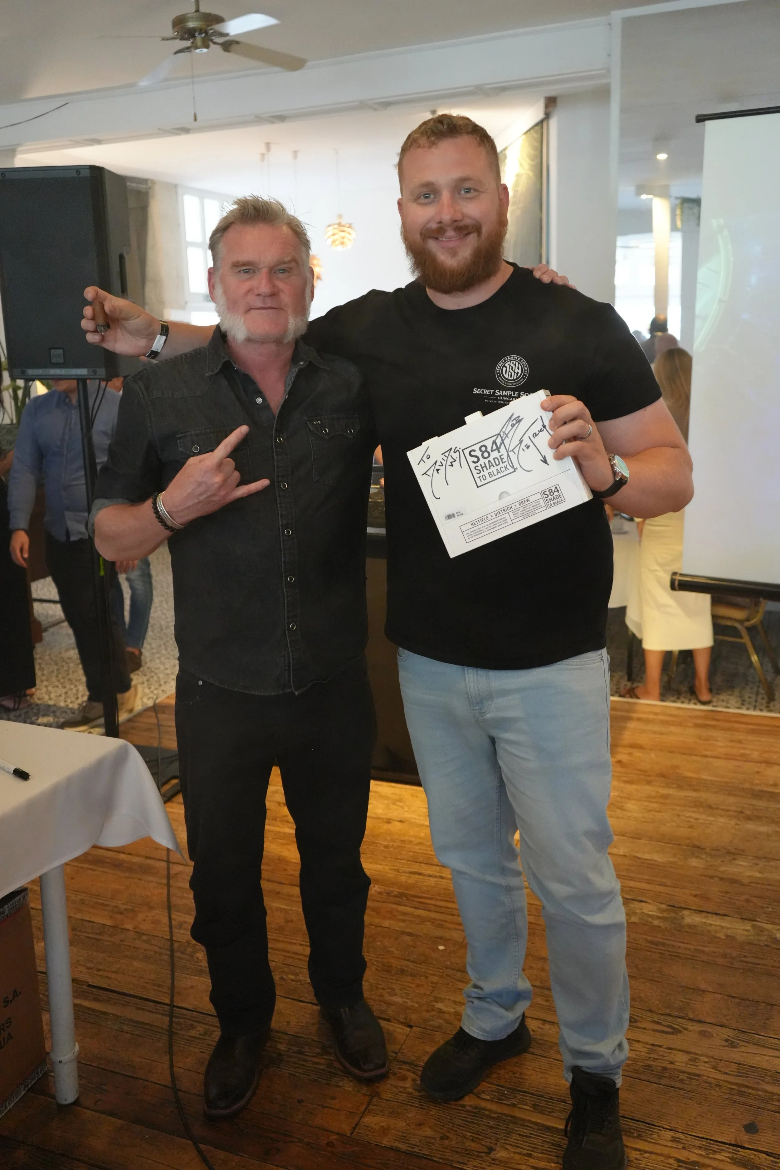 Two men standing together inside a room, smiling at the camera. The man on the left has gray hair and a beard, wearing a dark button-up shirt. The man on the right has short red hair and a beard, wearing a black T-shirt with writing on it, and holdin