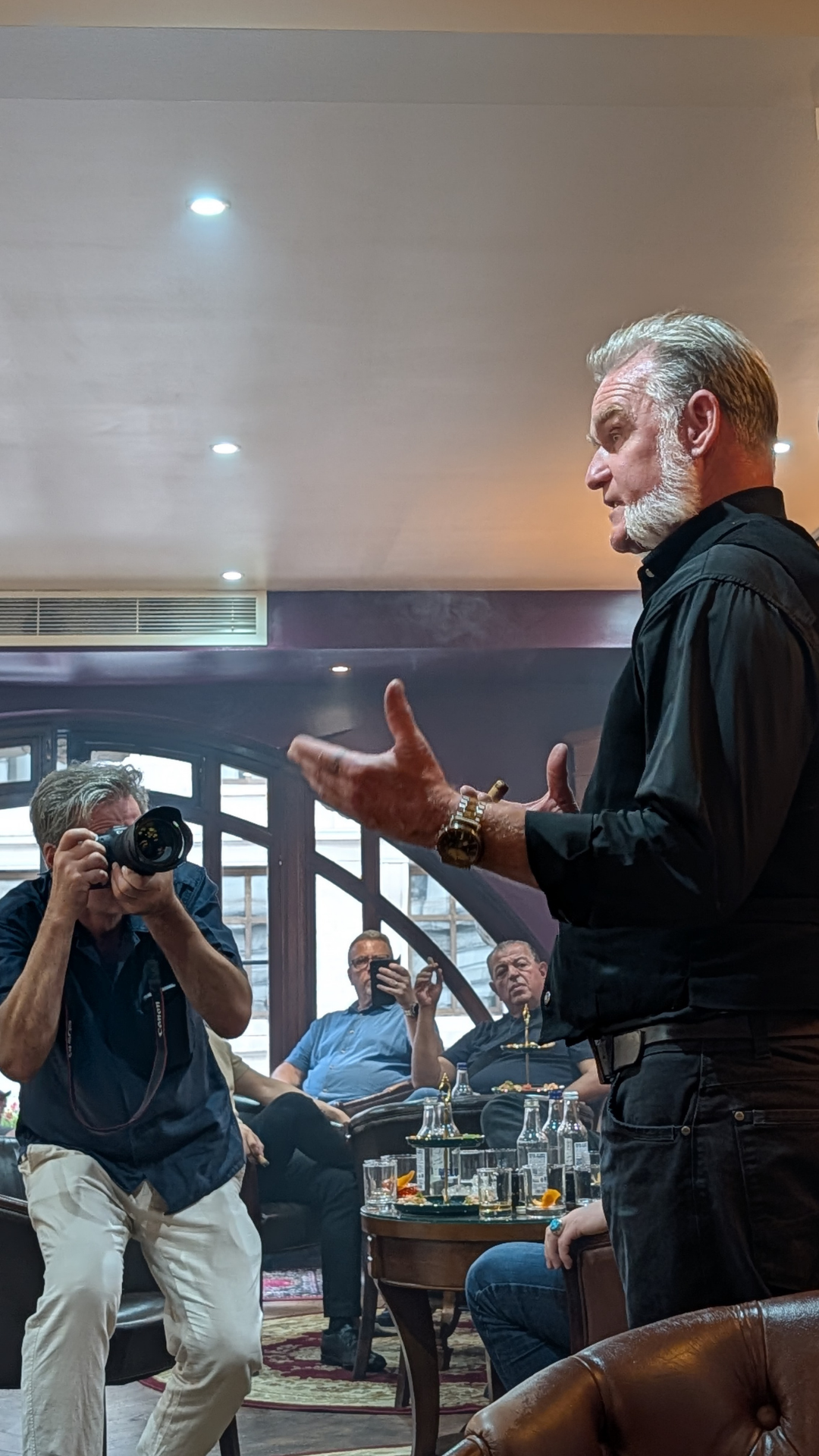 An older man with gray hair and beard, dressed in black, speaking to a group indoors while others listen and take photos of him.