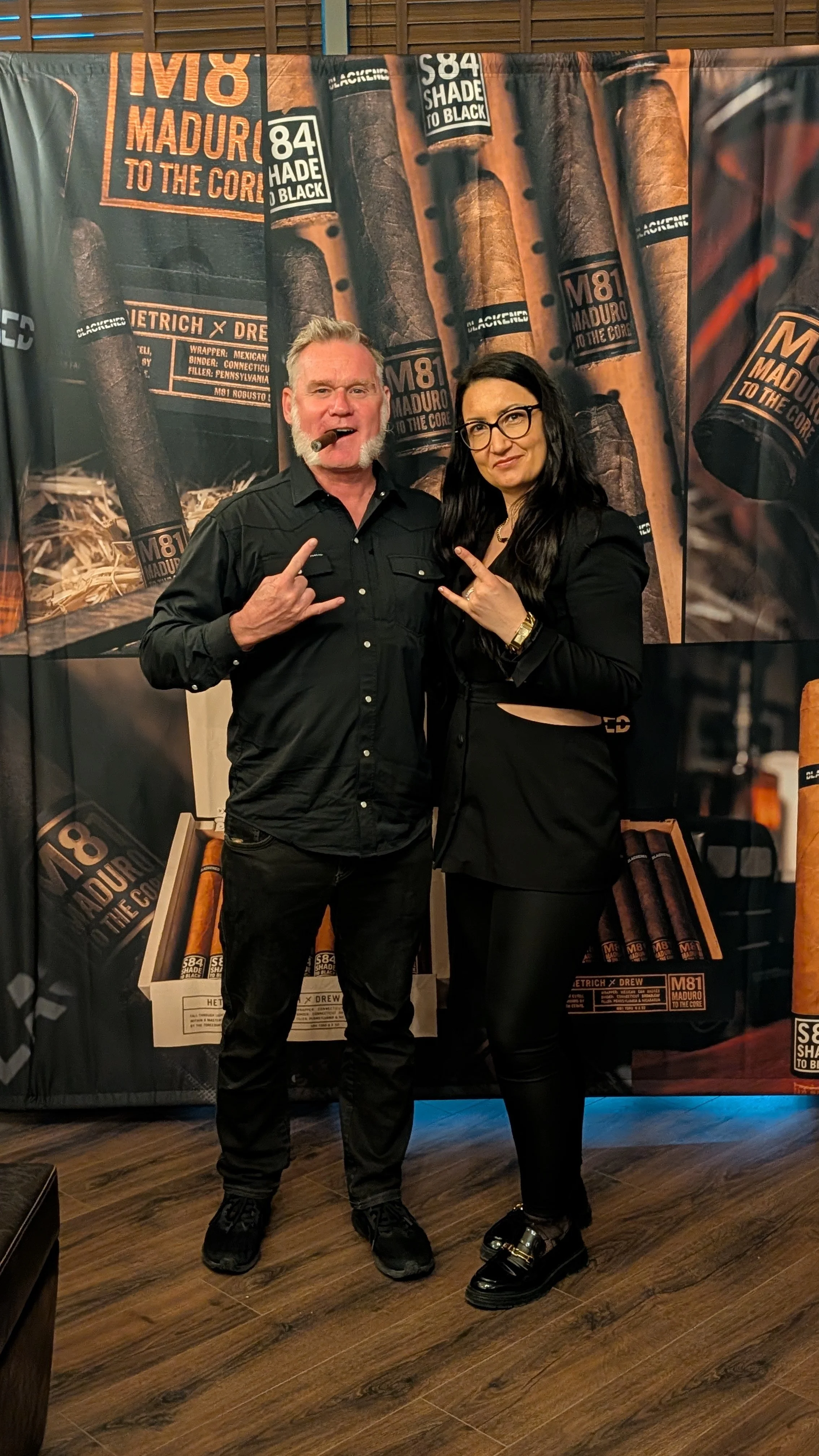 Two people standing in front of a backdrop with cigar boxes, making rock hand gestures.