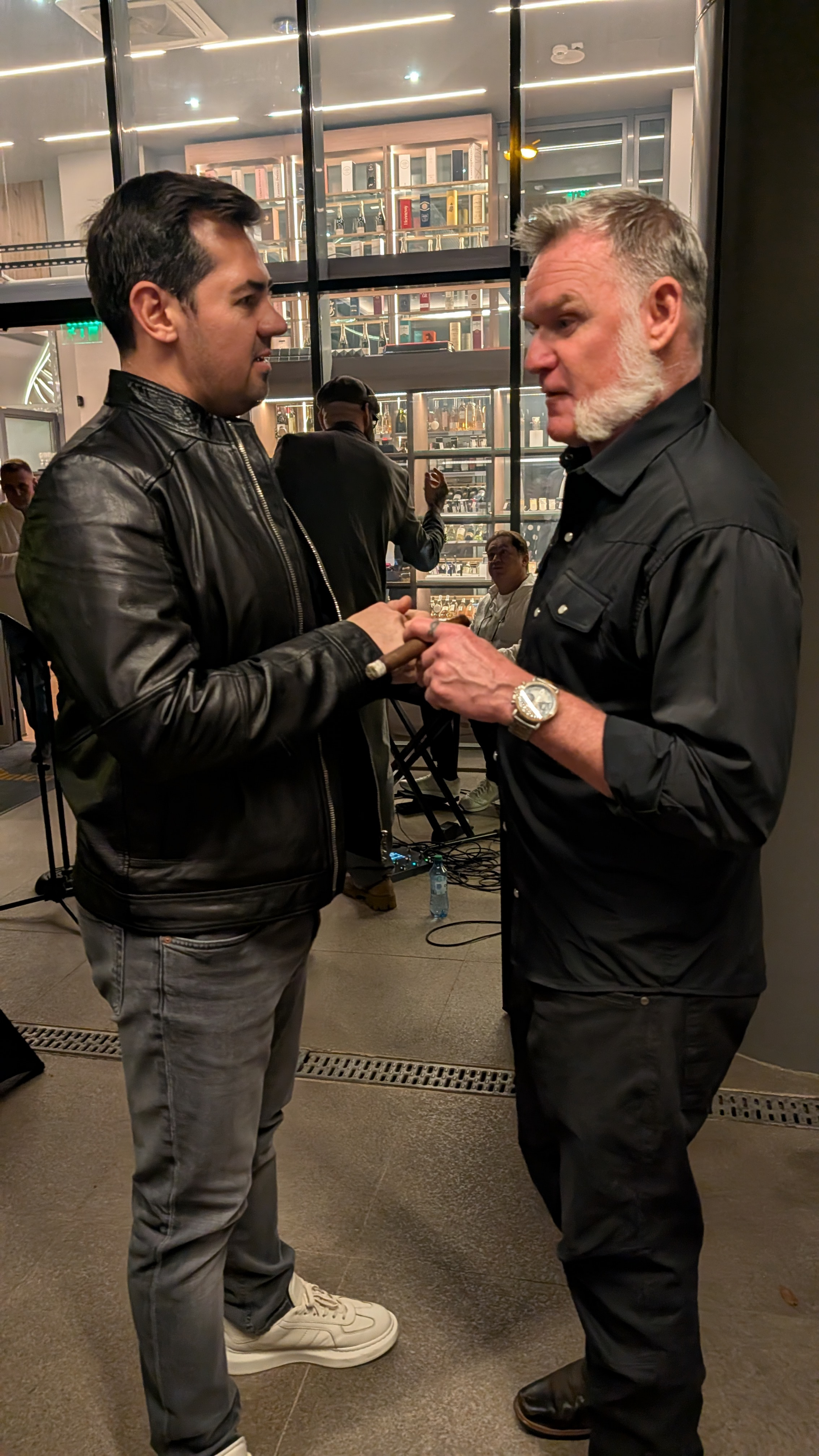 Two men are engaged in a conversation indoors, holding hands or a handshake, with a bar or liquor shelves in the background and other people nearby.