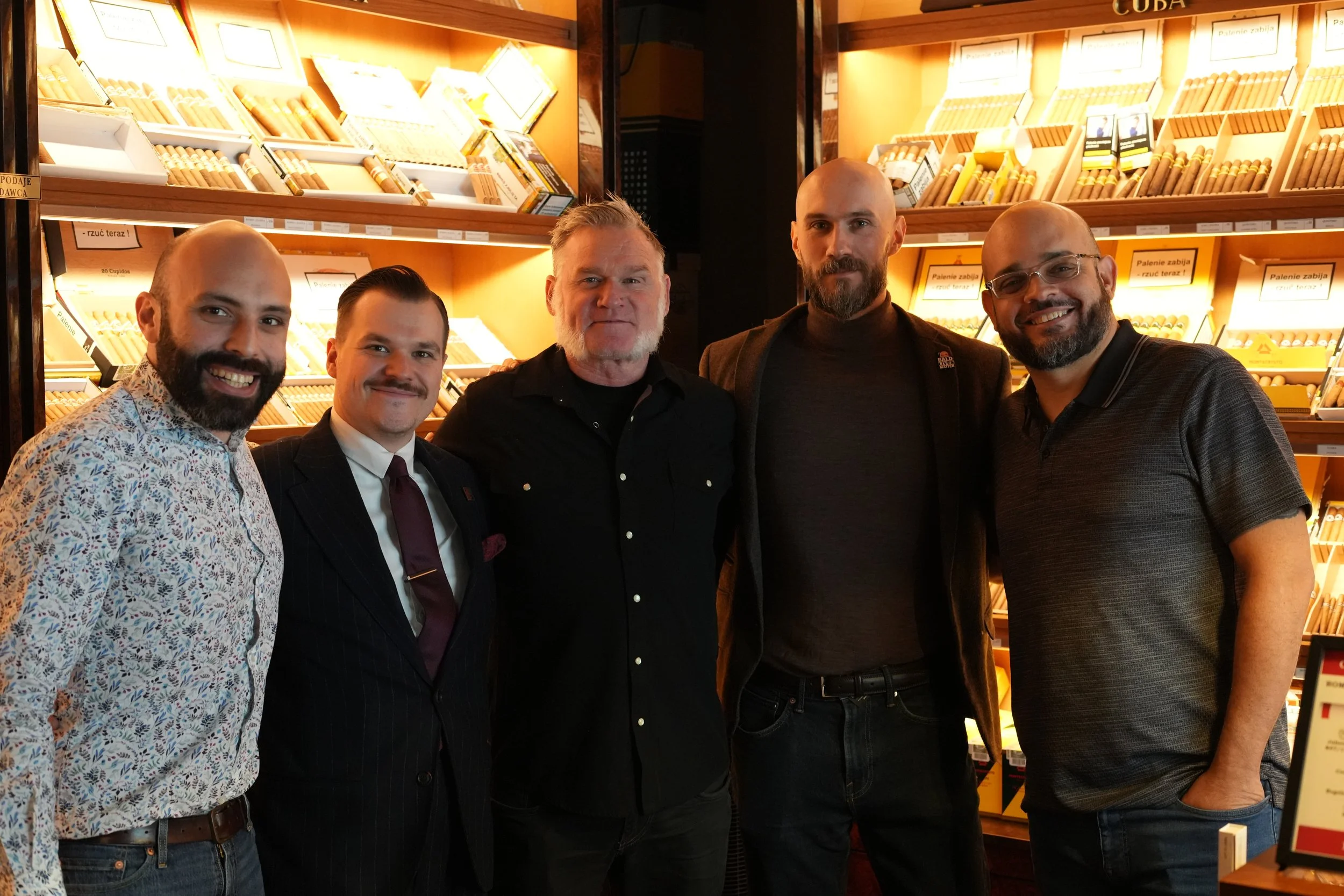 Five men dressed in business casual and formal attire standing in front of a cigar display shelf in a store, smiling and posing for the photo.