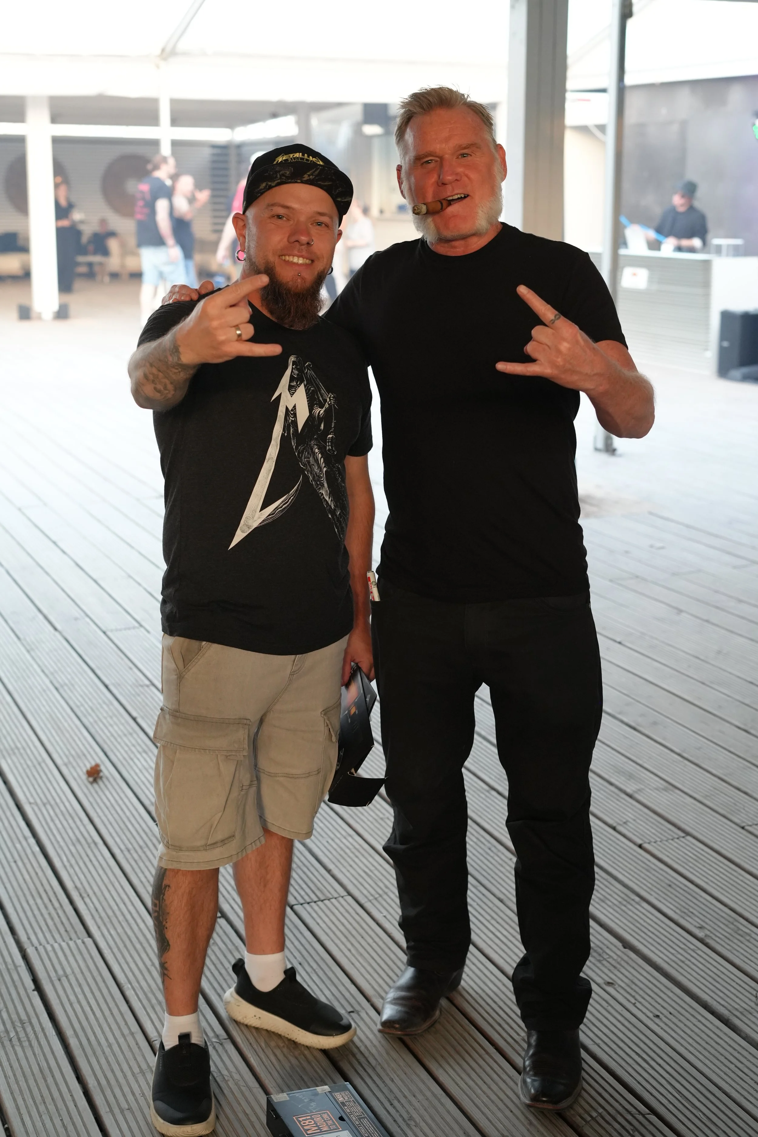 Two men standing side by side, smiling and making rock hand gestures. The man on the left is wearing a black t-shirt with a graphic, beige cargo shorts, a black cap, and has tattoos and piercings. The man on the right is older, with gray hair and a b