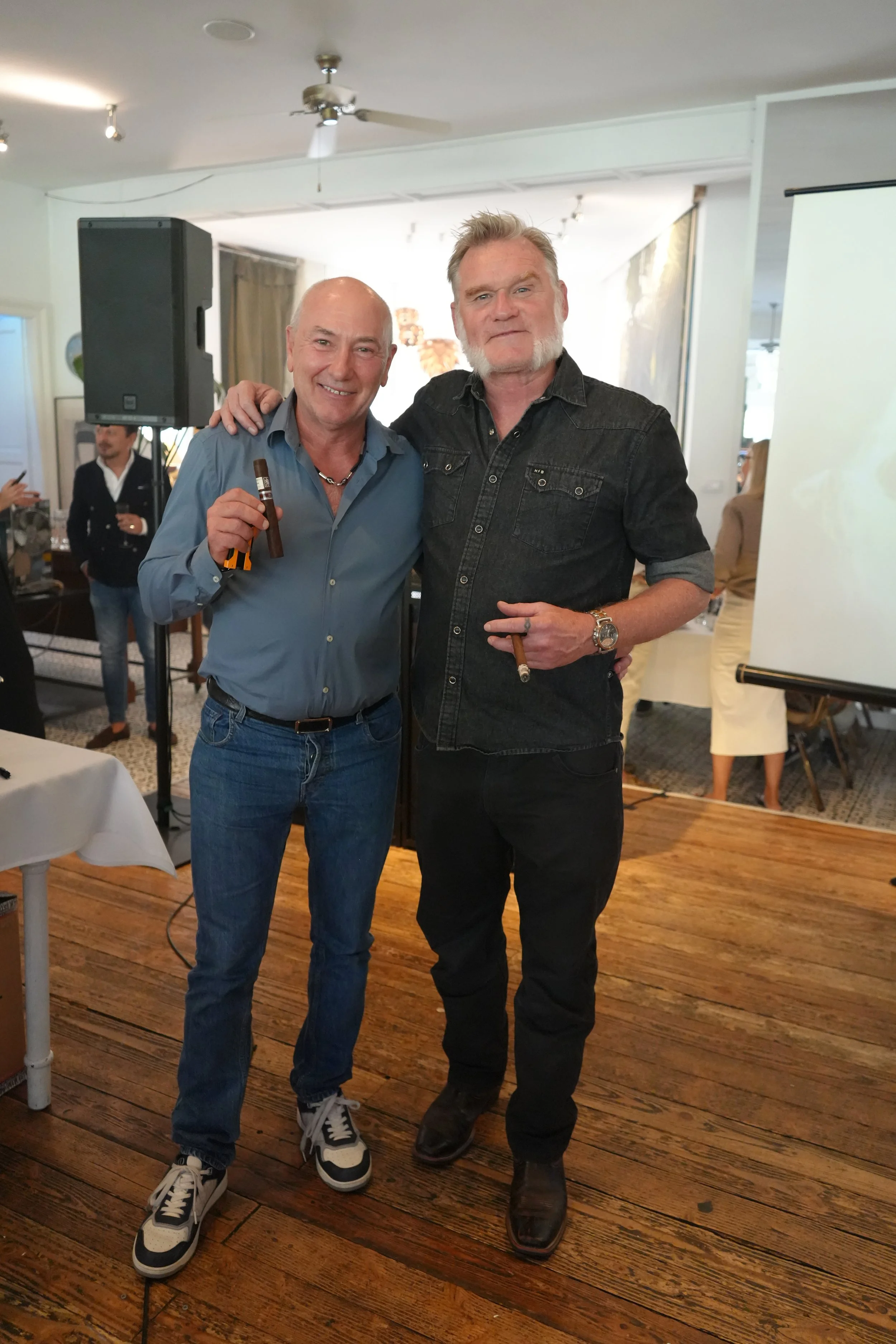 Two men standing closely together, smiling, each holding a cigar in their hand, in an indoor venue with wooden floors, a white projection screen, and other people in the background.