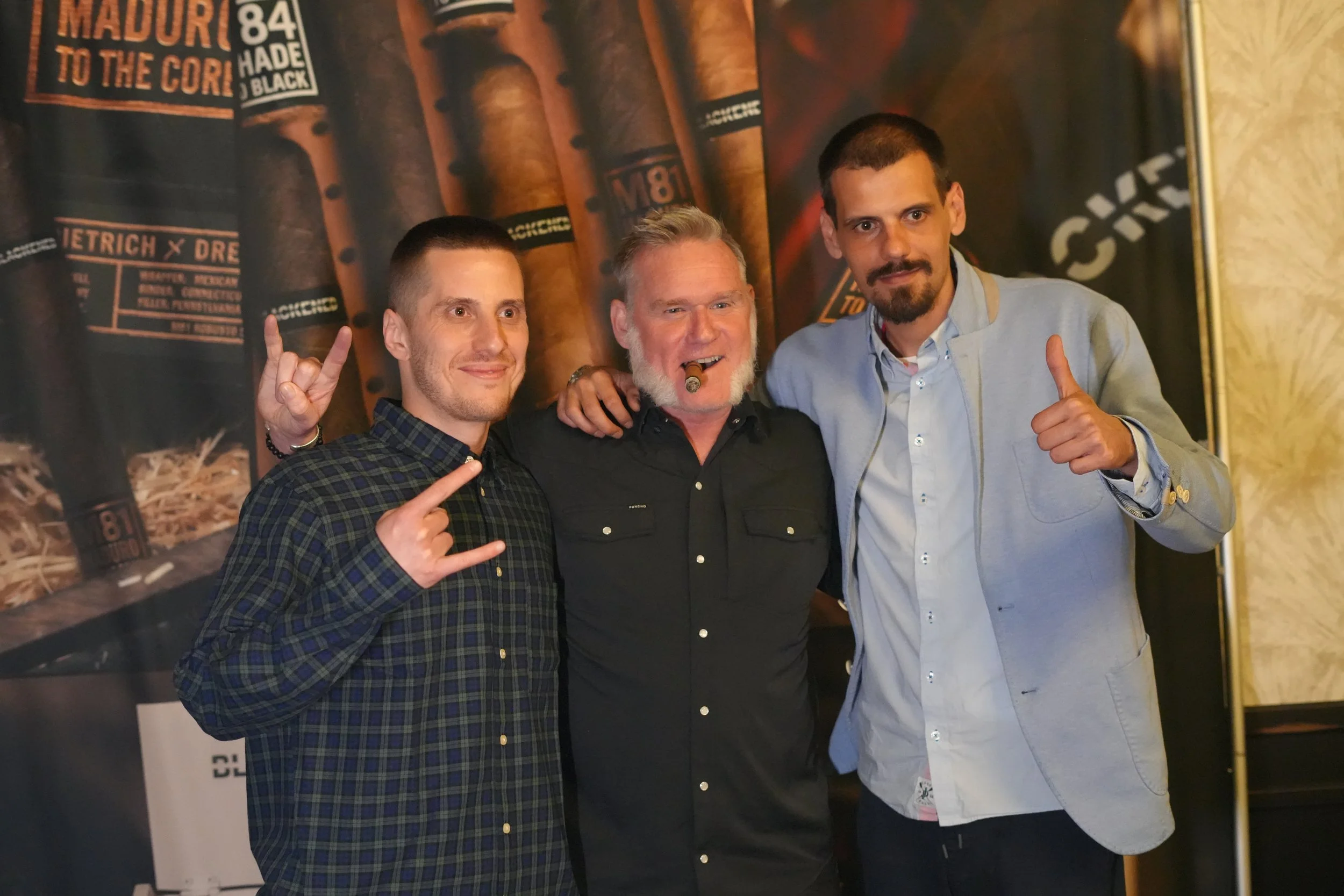 Three men standing together, smiling, and making hand gestures. The man on the left is wearing a plaid shirt and making a rock on sign with his fingers. The man in the middle, who has gray hair and a beard, is holding a cigar and making a peace sign.