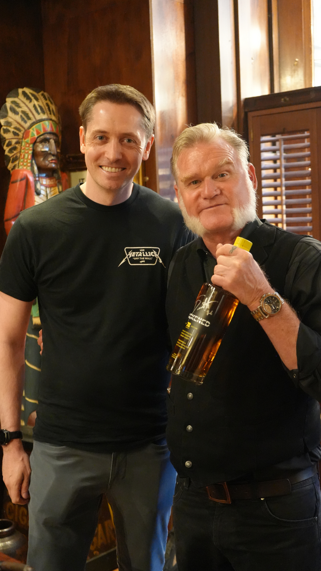 Two men standing together, one holding a drink, inside a wooden-paneled room with a decorative African mask in the background.