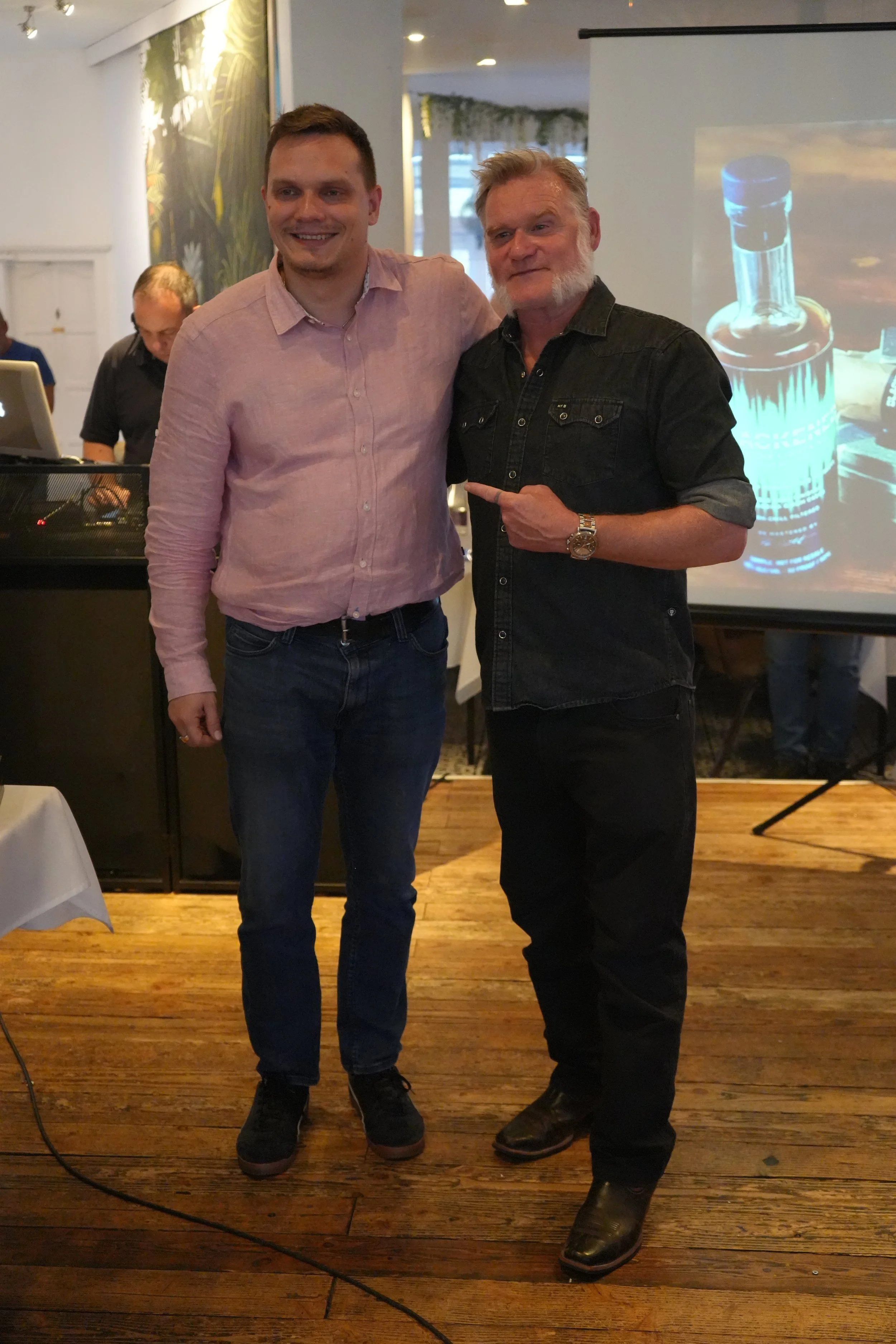 Two men standing side by side in a room, smiling at the camera. They are indoors with wooden flooring and a large projection screen displaying an image of a glass bottle. One man is wearing a pink button-up shirt and jeans, and the other is dressed i