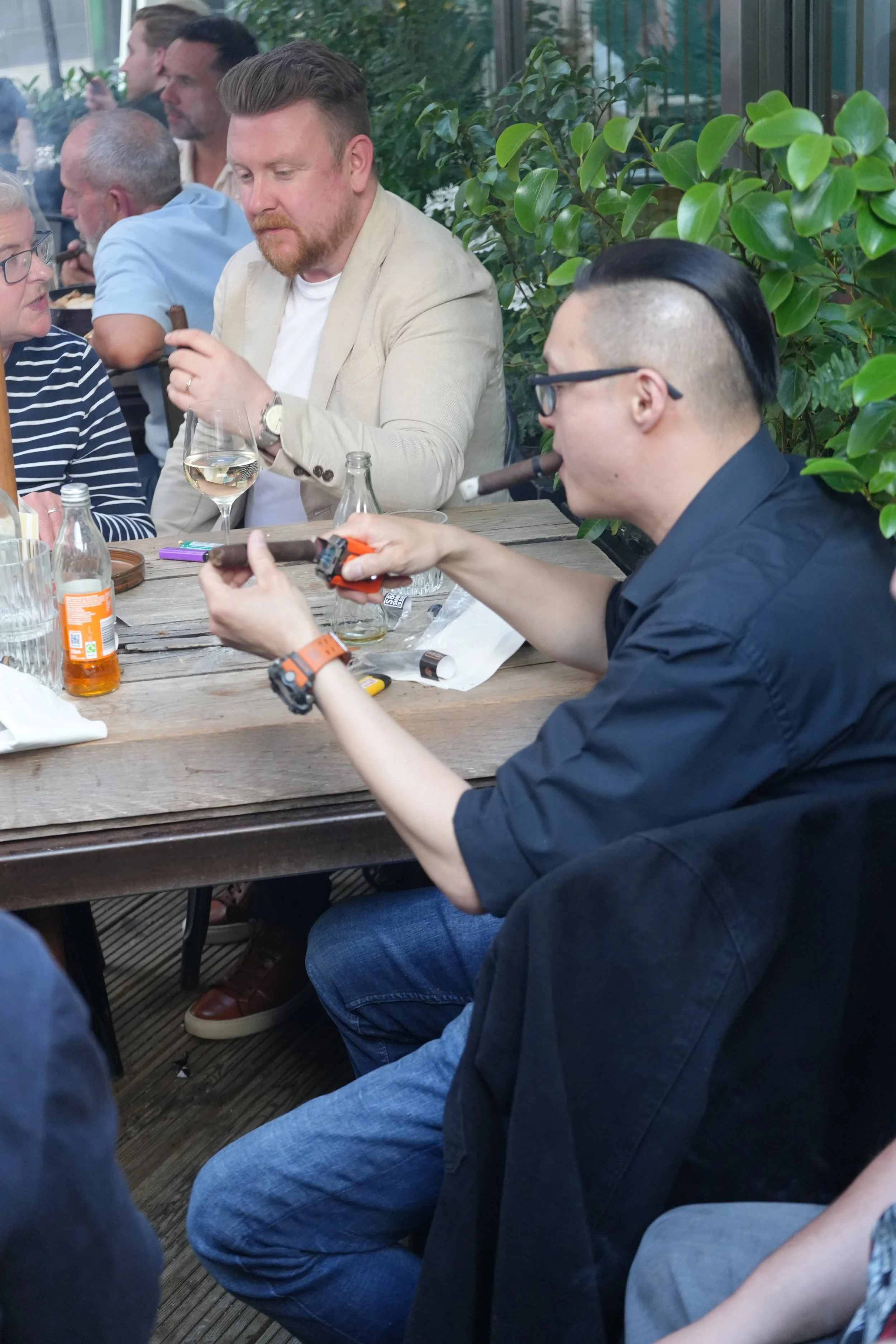 People sitting at a wooden table in a greenhouse-like setting, some smoking cigars, with greenery in the background, and various bottles and glasses on the table.