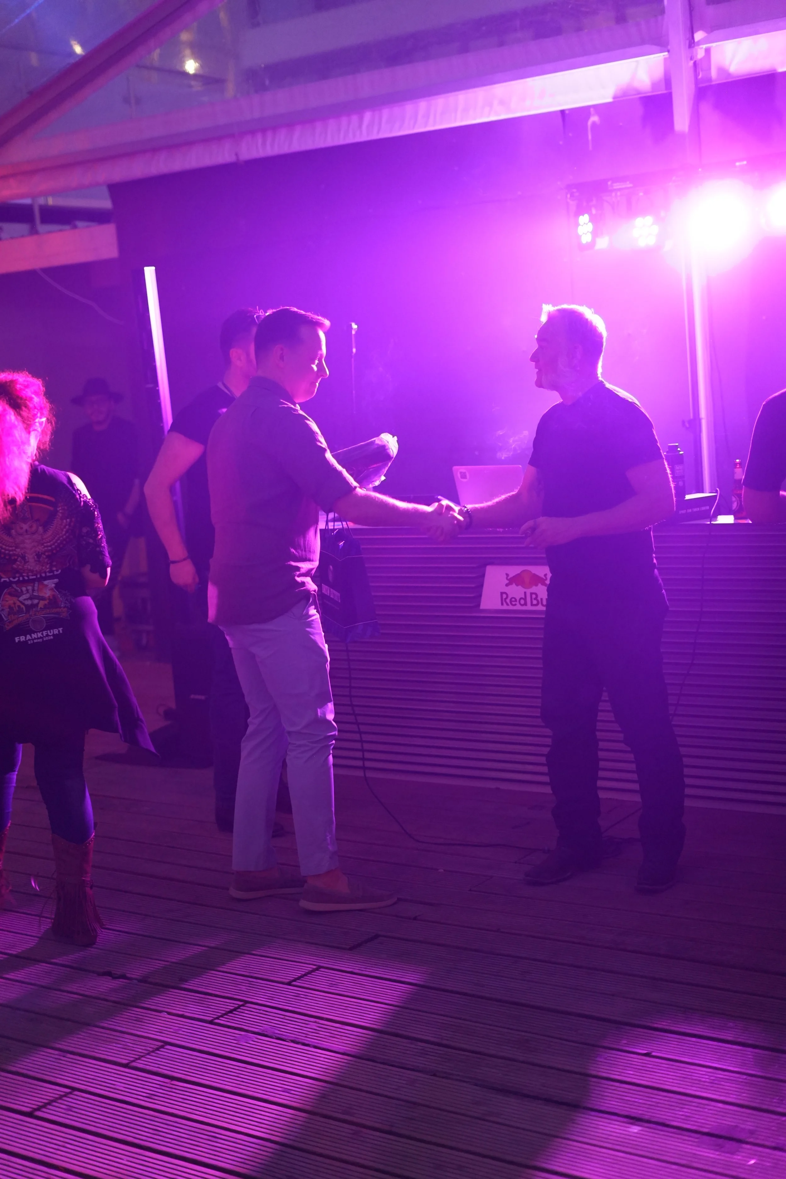 Two men shaking hands in a purple-lit nightclub or concert venue with a DJ booth in the background.