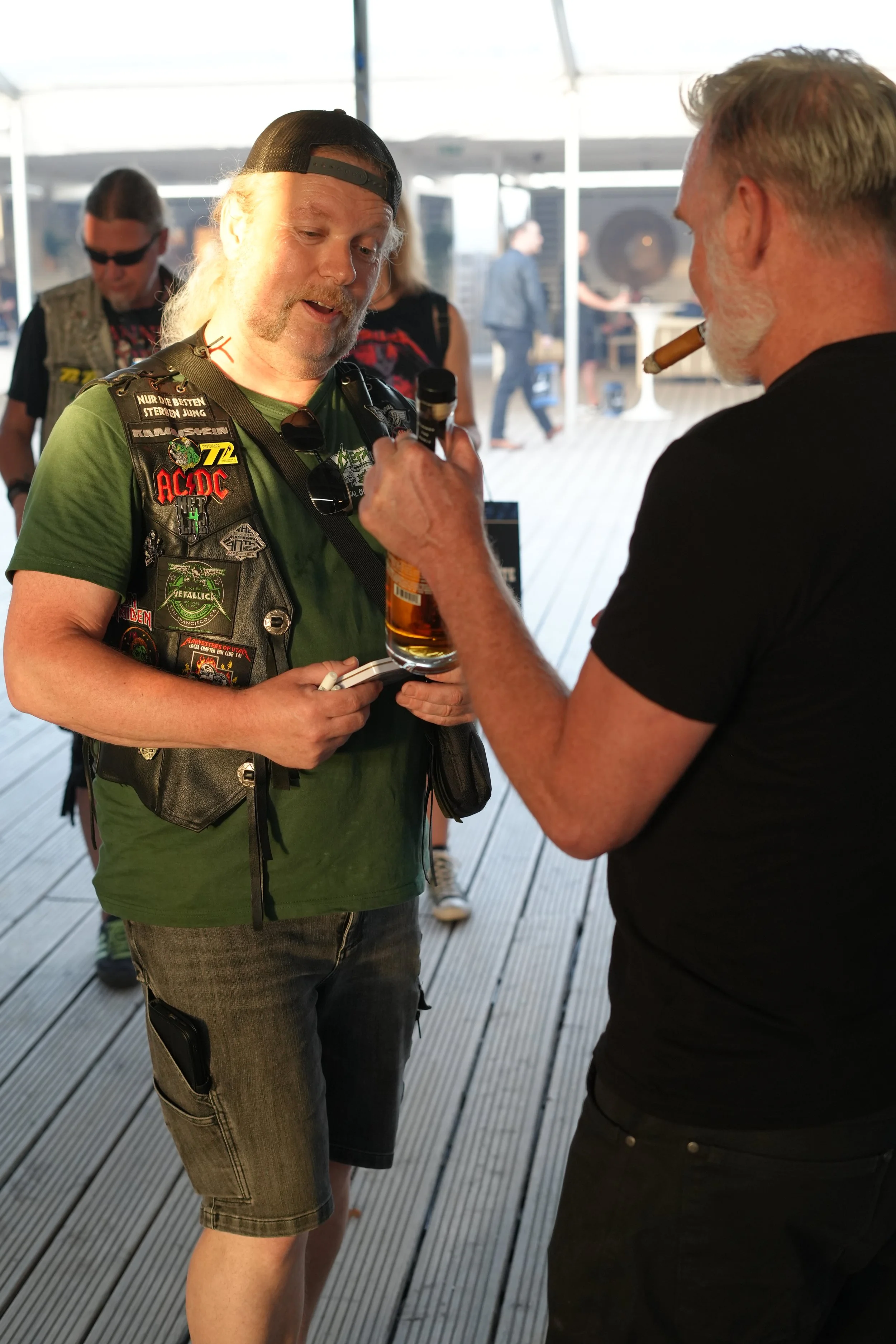 Two men having a conversation inside a tented area, one holding a microphone and a beer, the other smoking a cigar. Several other people are visible in the background.