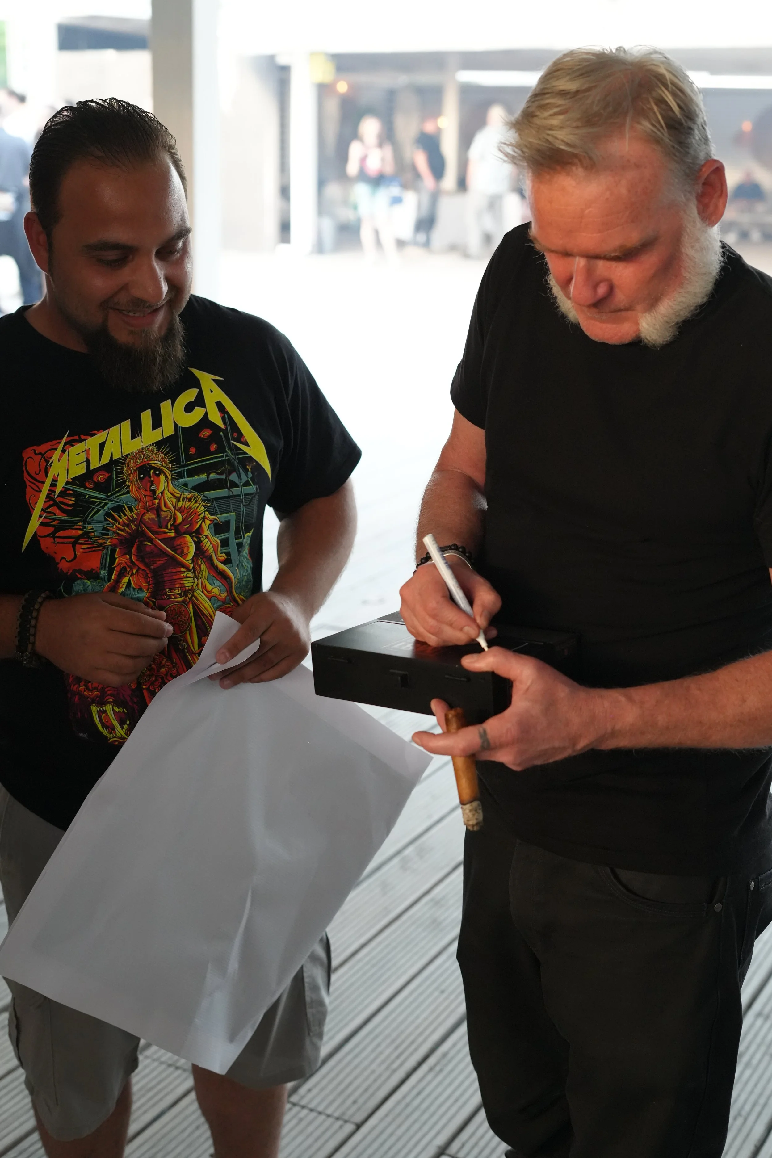 Two men signing papers or a book at an outdoor event, with a bright background and some blurred people in the distance.