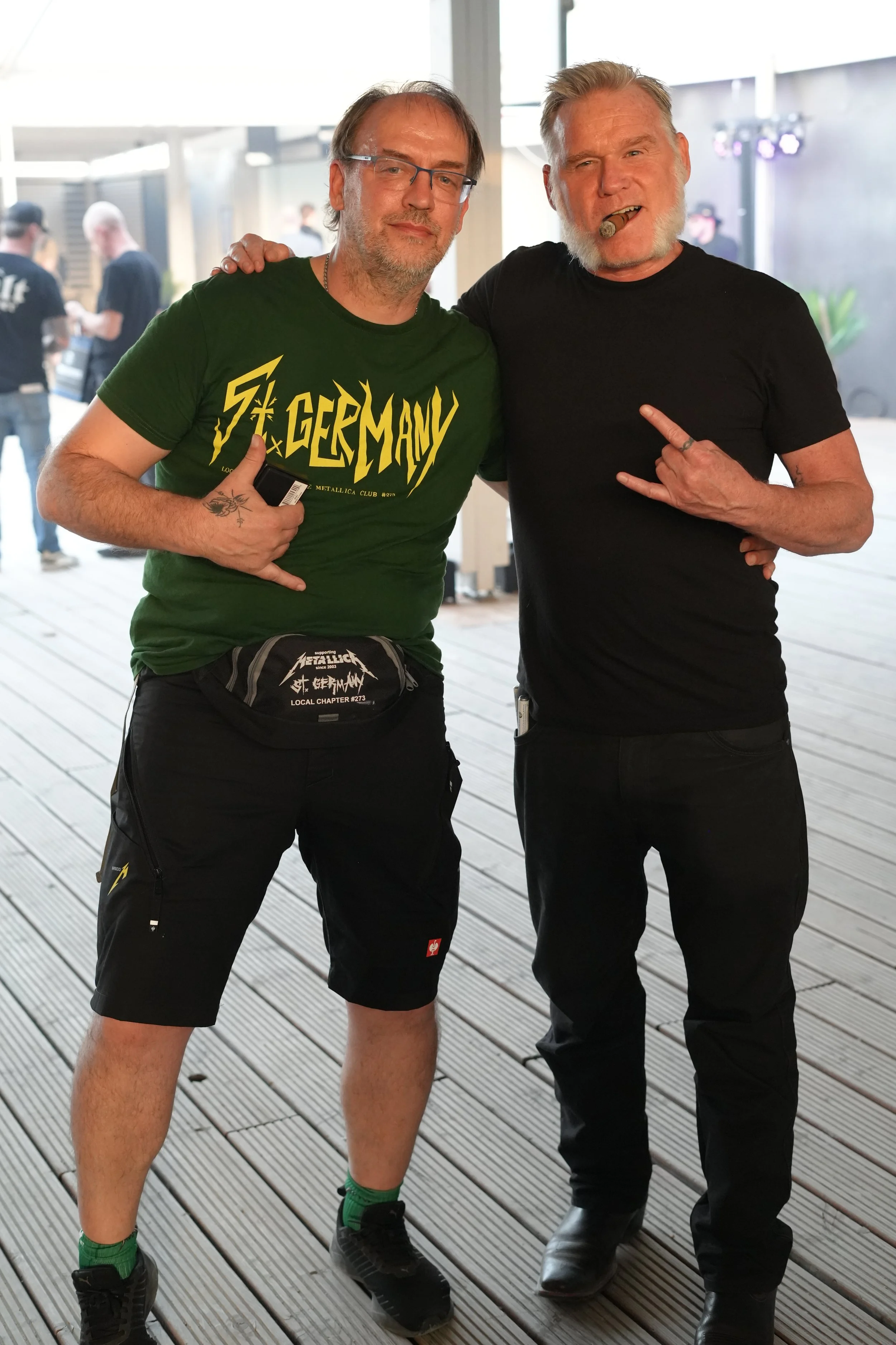 Two men standing together on a wooden deck, smiling, with one man wearing a green 'Germoney' t-shirt and the other wearing a black t-shirt and smoking a cigar.