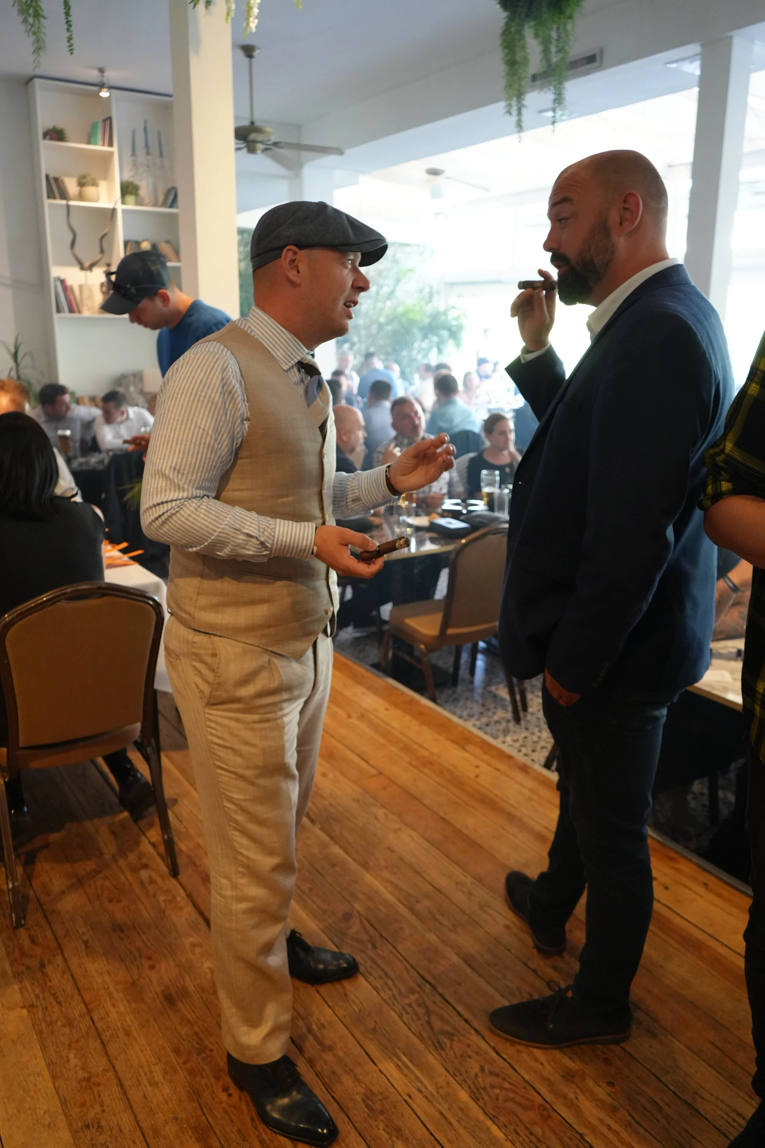 Two men are having a conversation at a social gathering inside a well-lit room. One man is wearing a flat cap, striped shirt, beige vest, and beige pants, holding a cigar. The other man has a shaved head, beard, and is dressed in a navy suit, holding