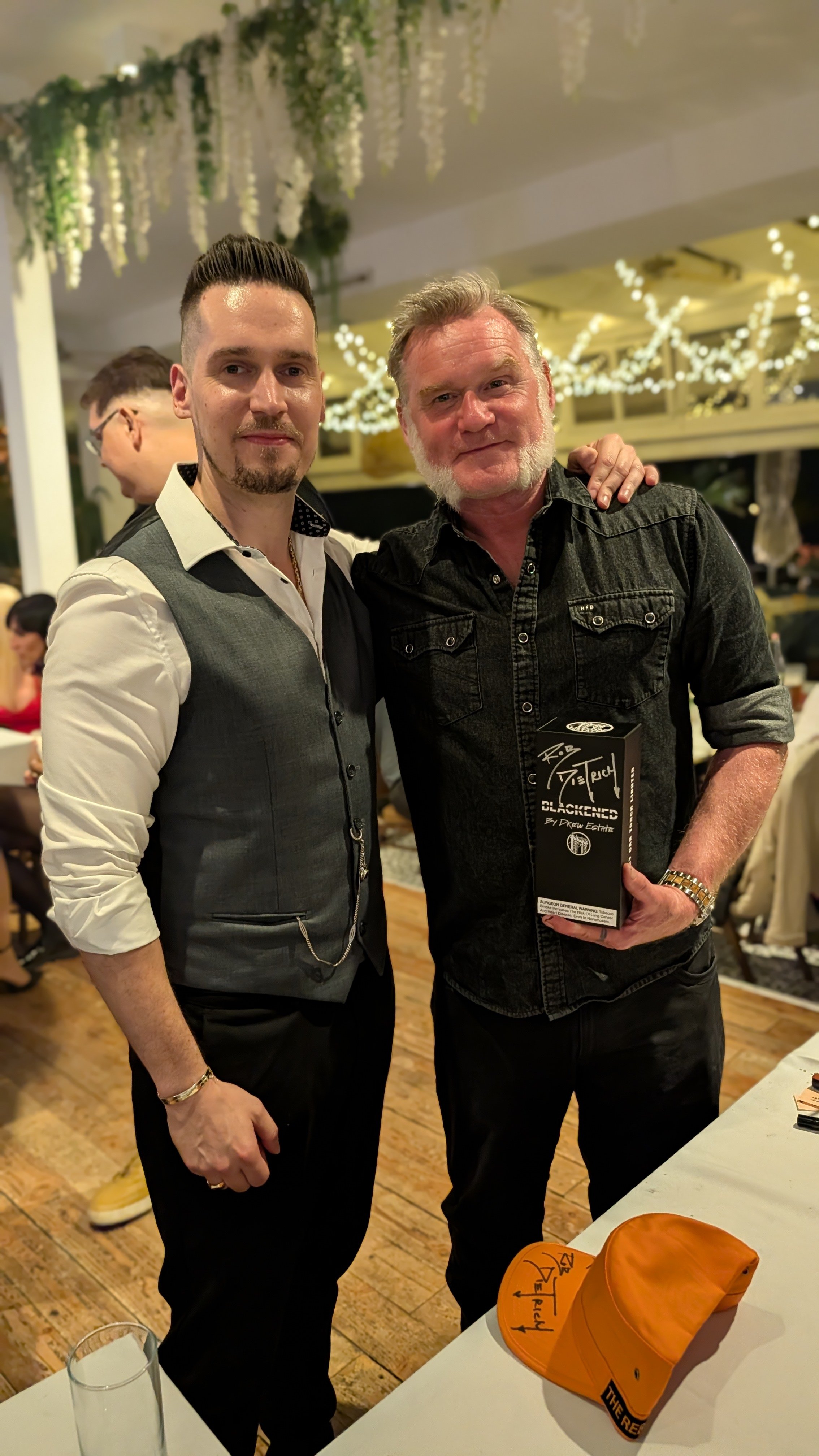 Two men standing together at an indoor event, one holding a black award or box, with decorative string lights and other guests in the background.