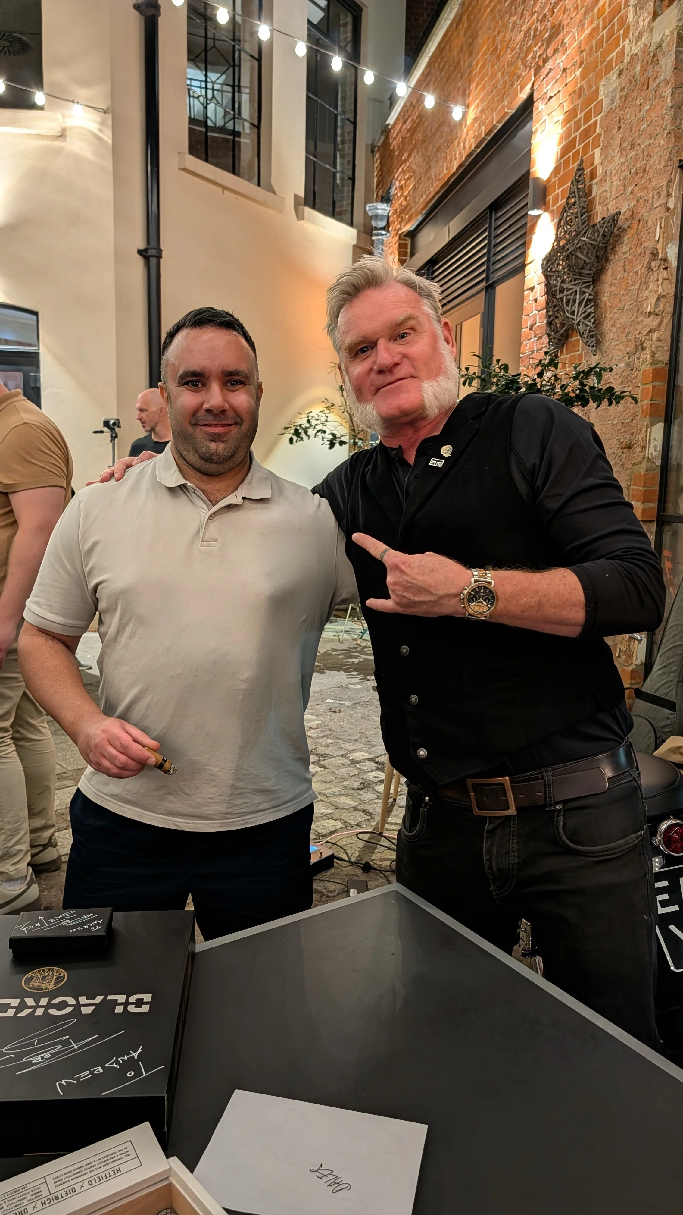 Two men standing together at an outdoor event in front of a brick wall with lighting and wall decorations. One man has short dark hair, a beard, and is wearing a light-colored polo shirt. The other man has gray hair, a beard, and is wearing a black s
