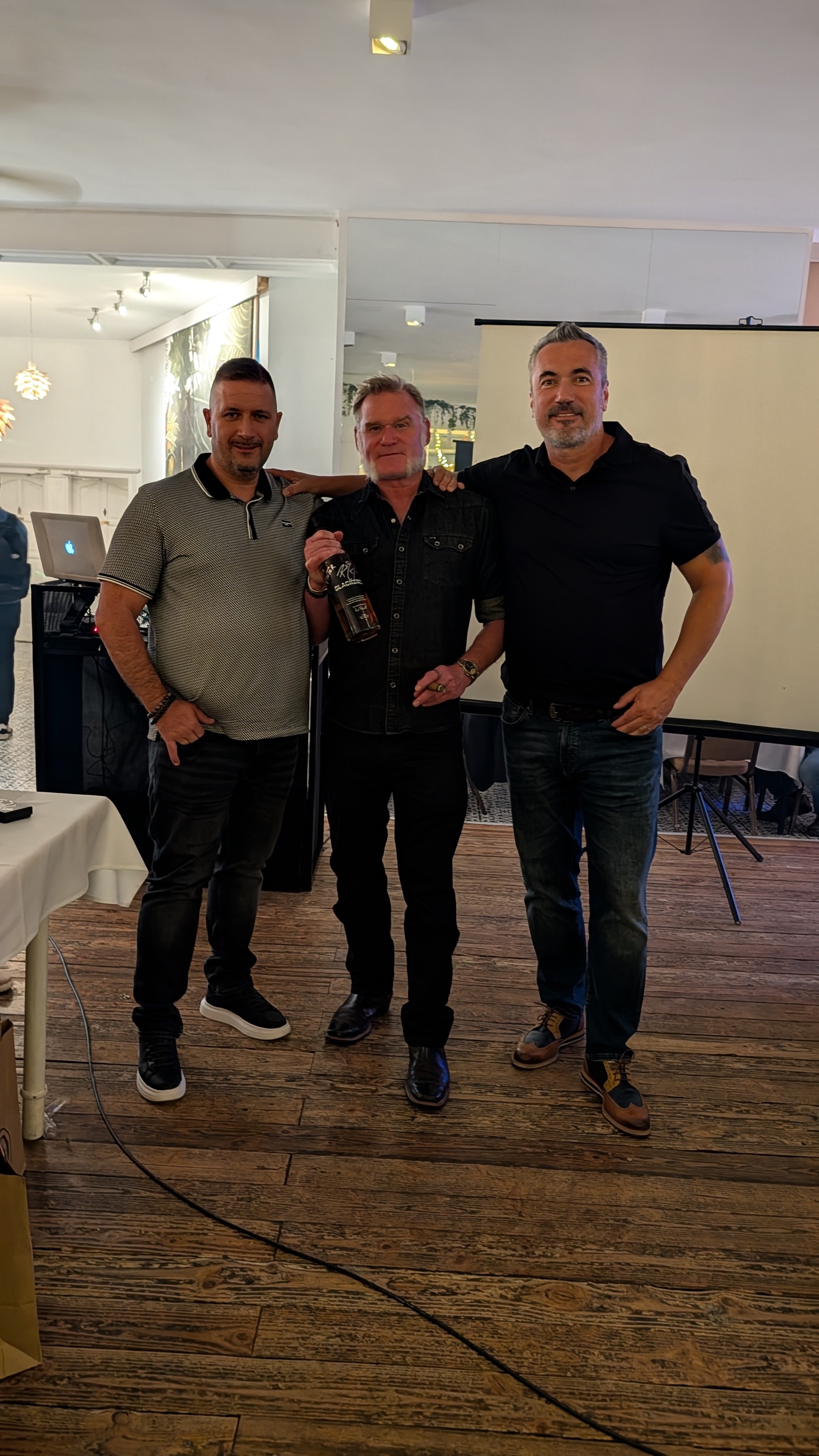 Three men standing together in a room, smiling for a photo. The man in the middle is holding a bottle of liquor.