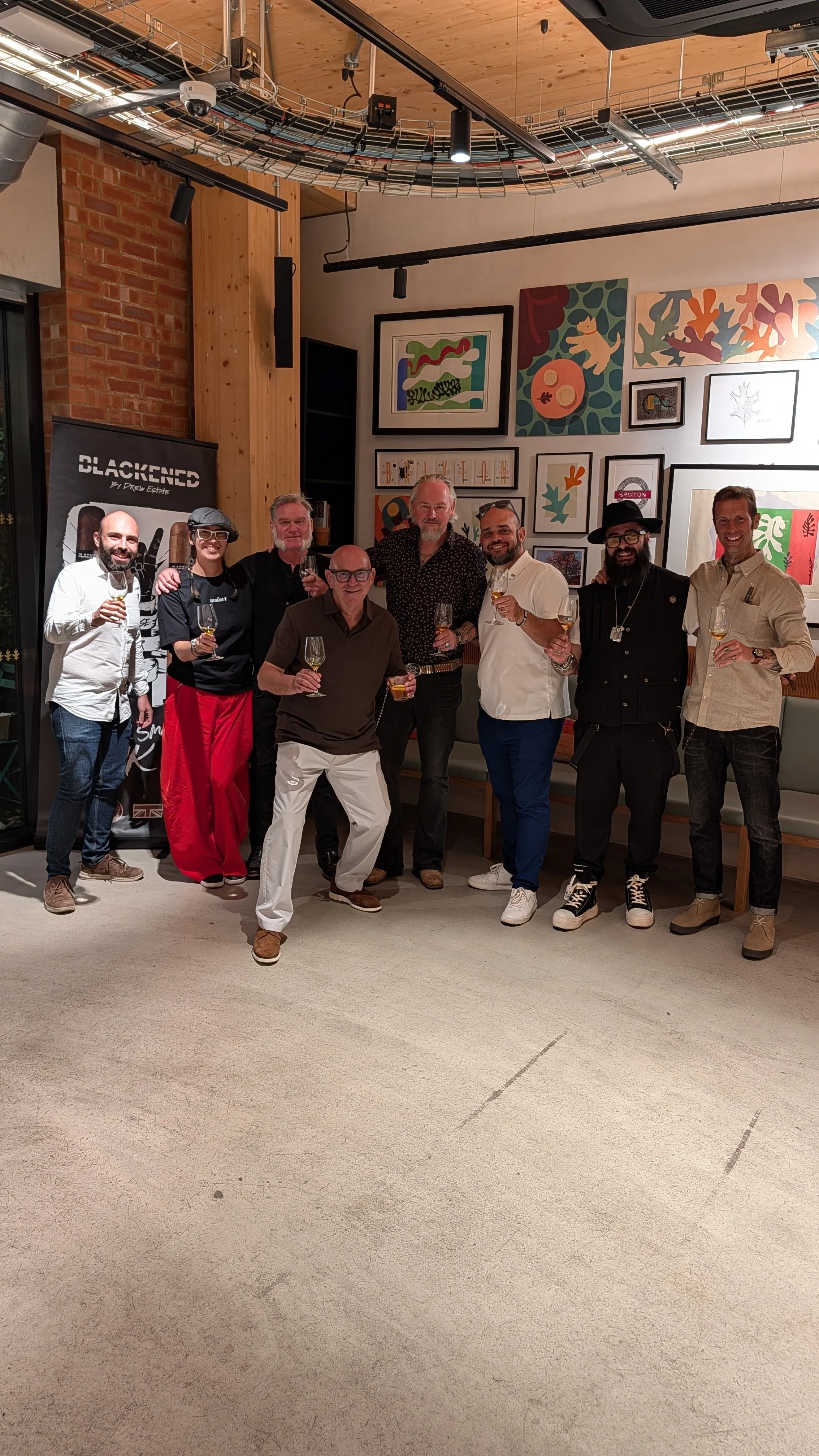 A group of eight people standing together in a room, holding glasses of wine, smiling, and posing for a photo. The room has a modern decor with framed artwork on the wall, and wooden and brick accents.
