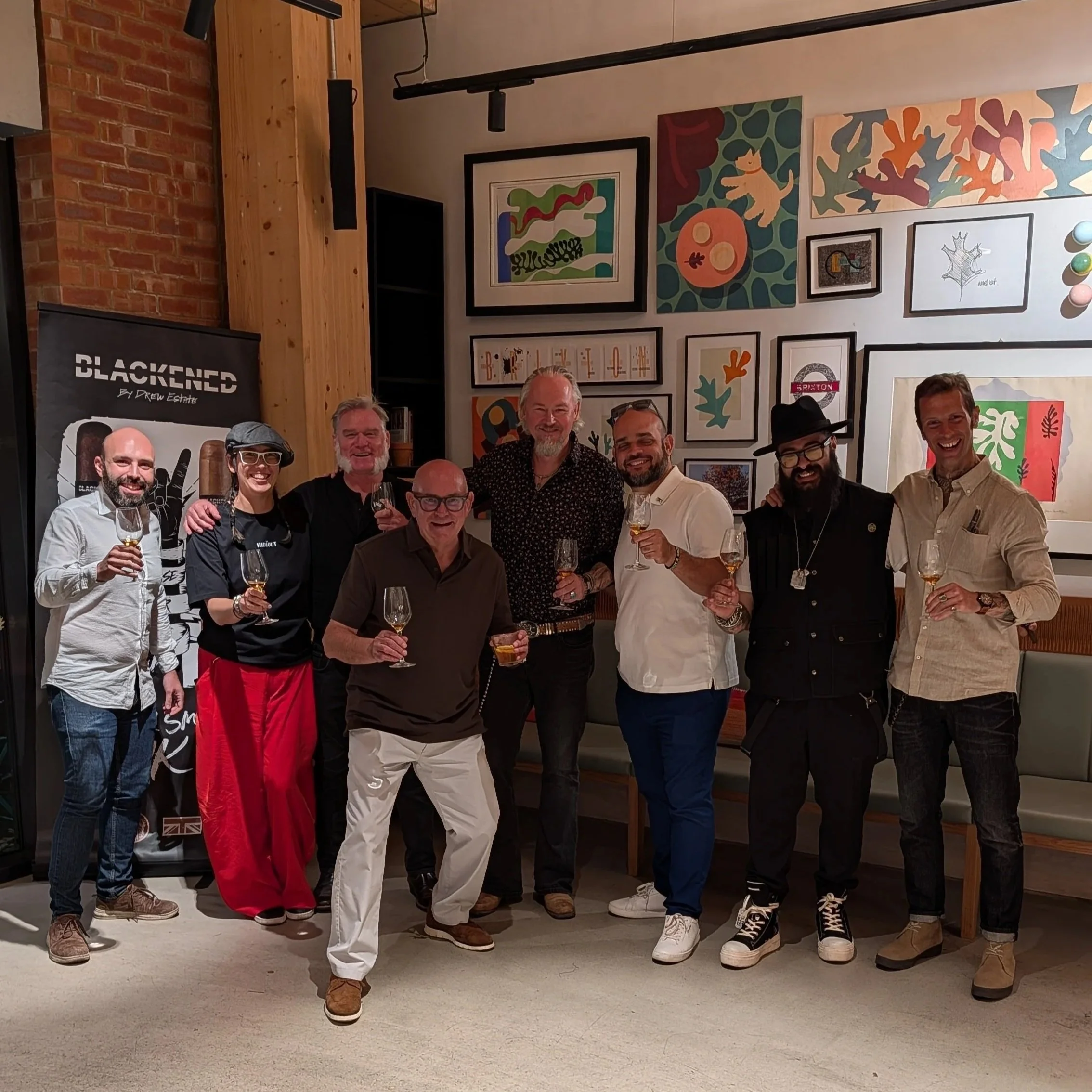 A group of people gathered together indoors, smiling and holding glasses of wine in a social setting, with colorful artwork on the wall behind them.