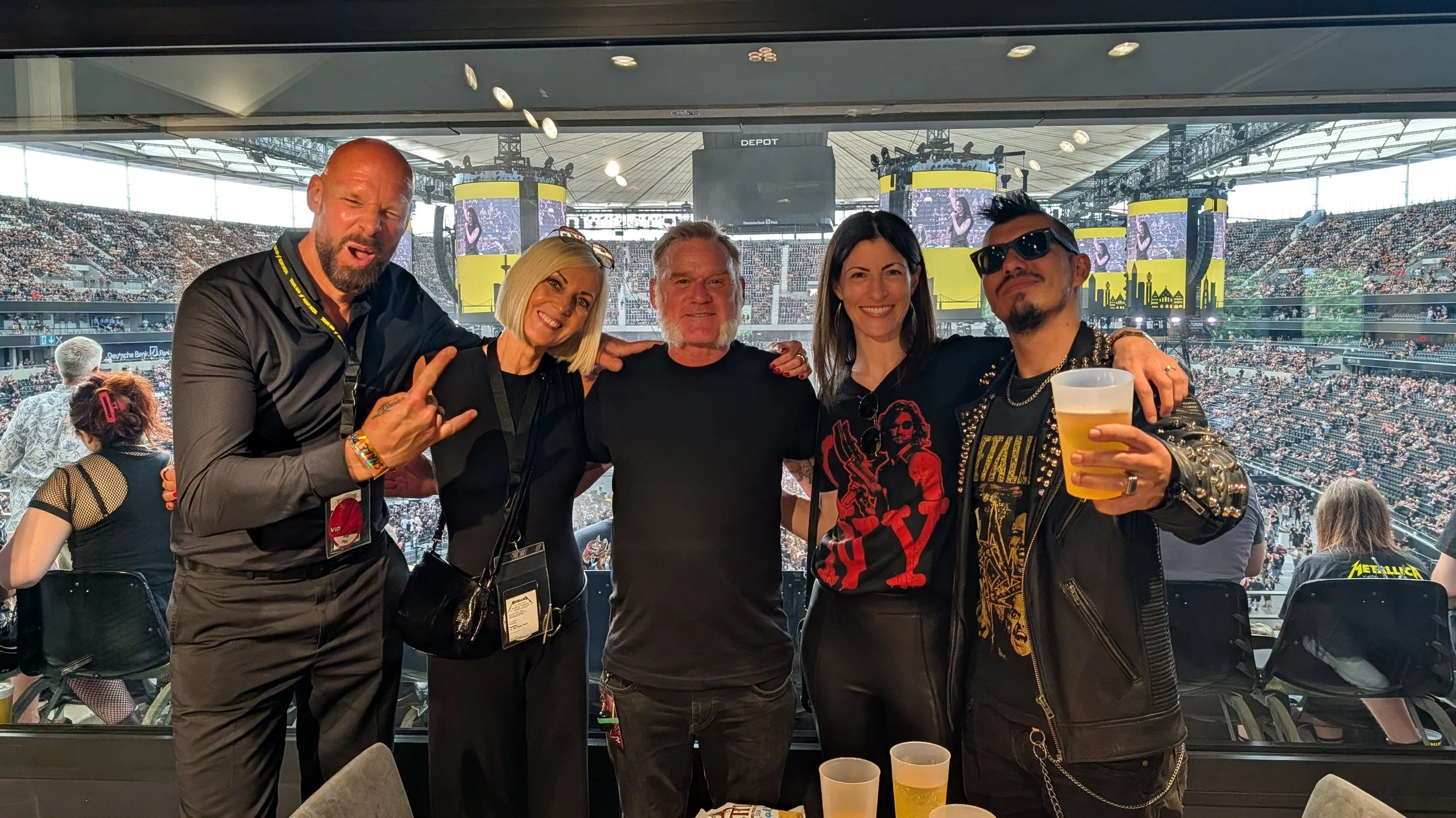 Group of five people posing for a photo at a large concert or event, with a packed stadium in the background and stage screens visible.