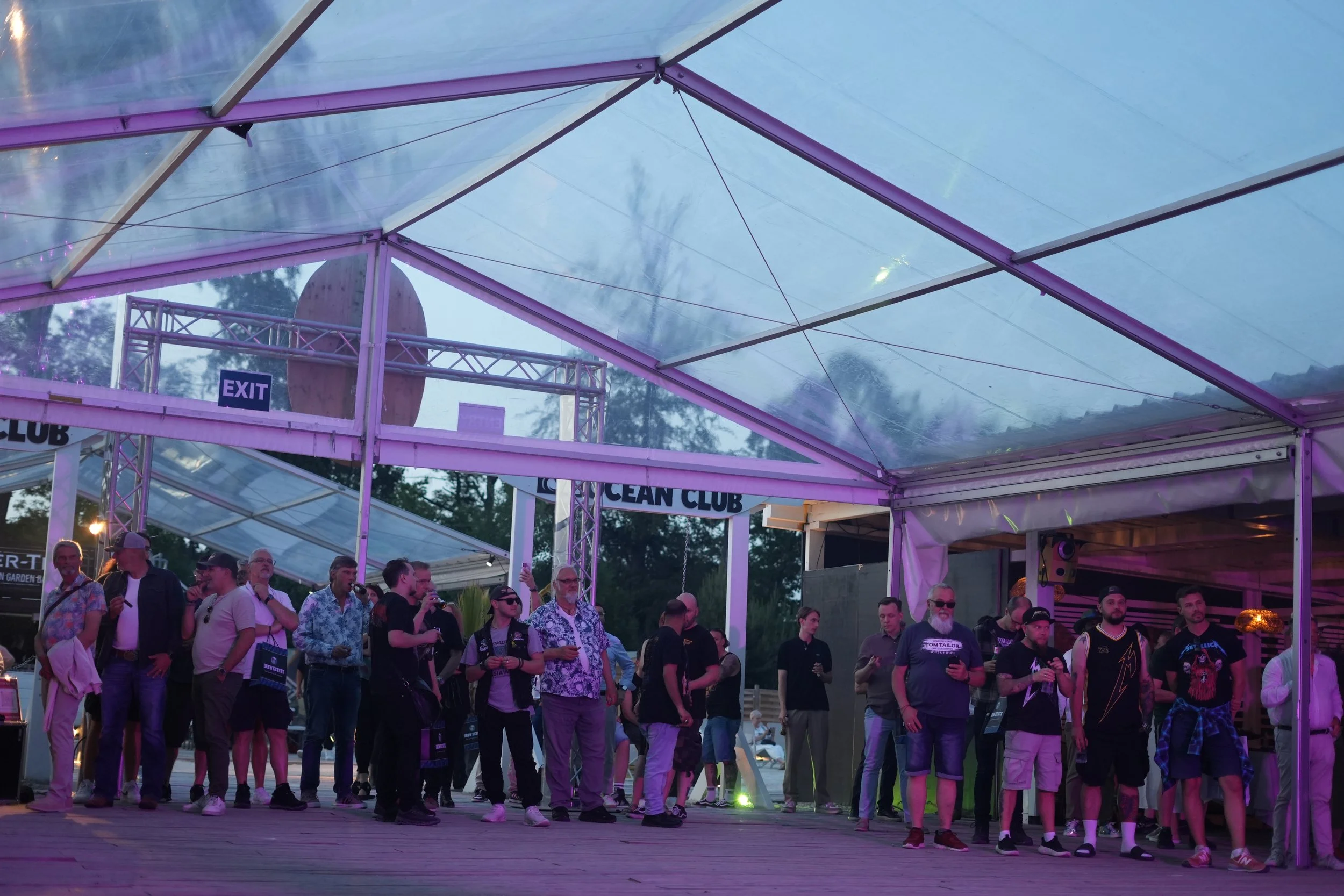 Crowd of people gathered at a music event under a large, transparent tent with purple lighting accents.