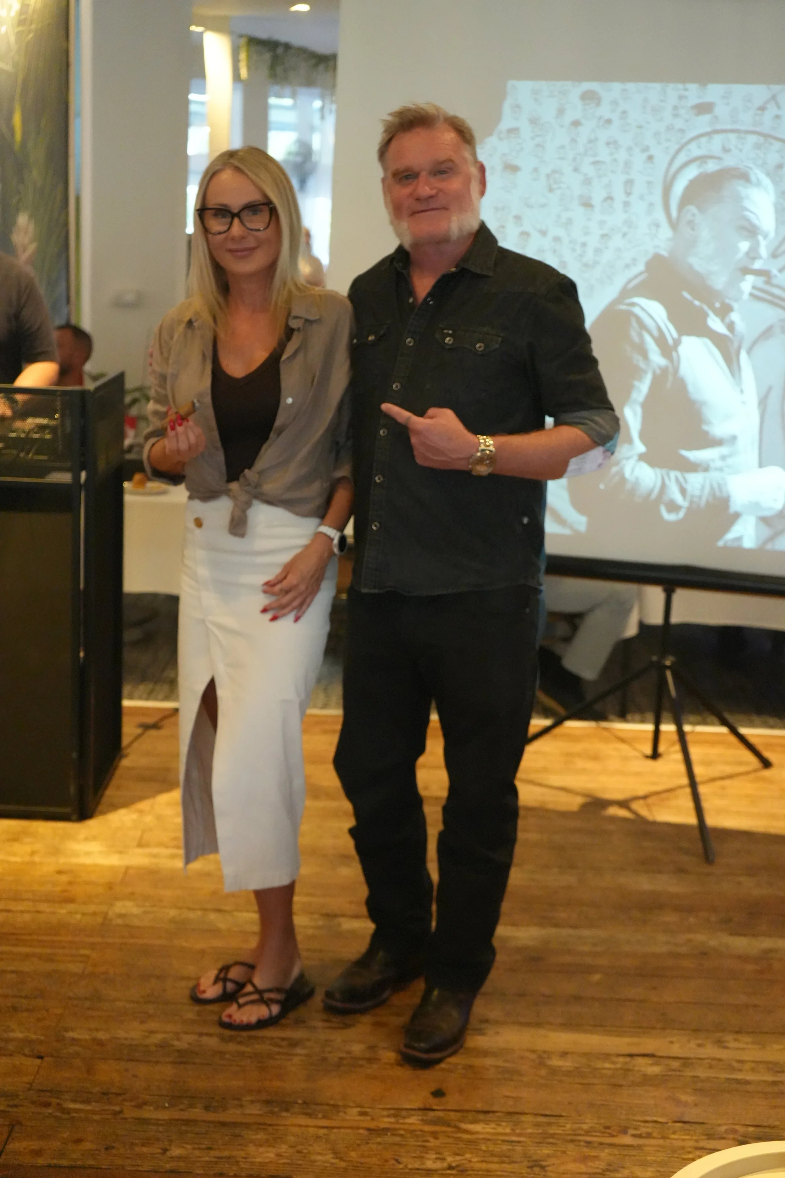 A woman with blonde hair, glasses, a beige shirt, and white skirt standing next to a man with blond hair, a beard, a black shirt, and black pants, pointing at him. They are indoors at an event with a projection and people in the background.