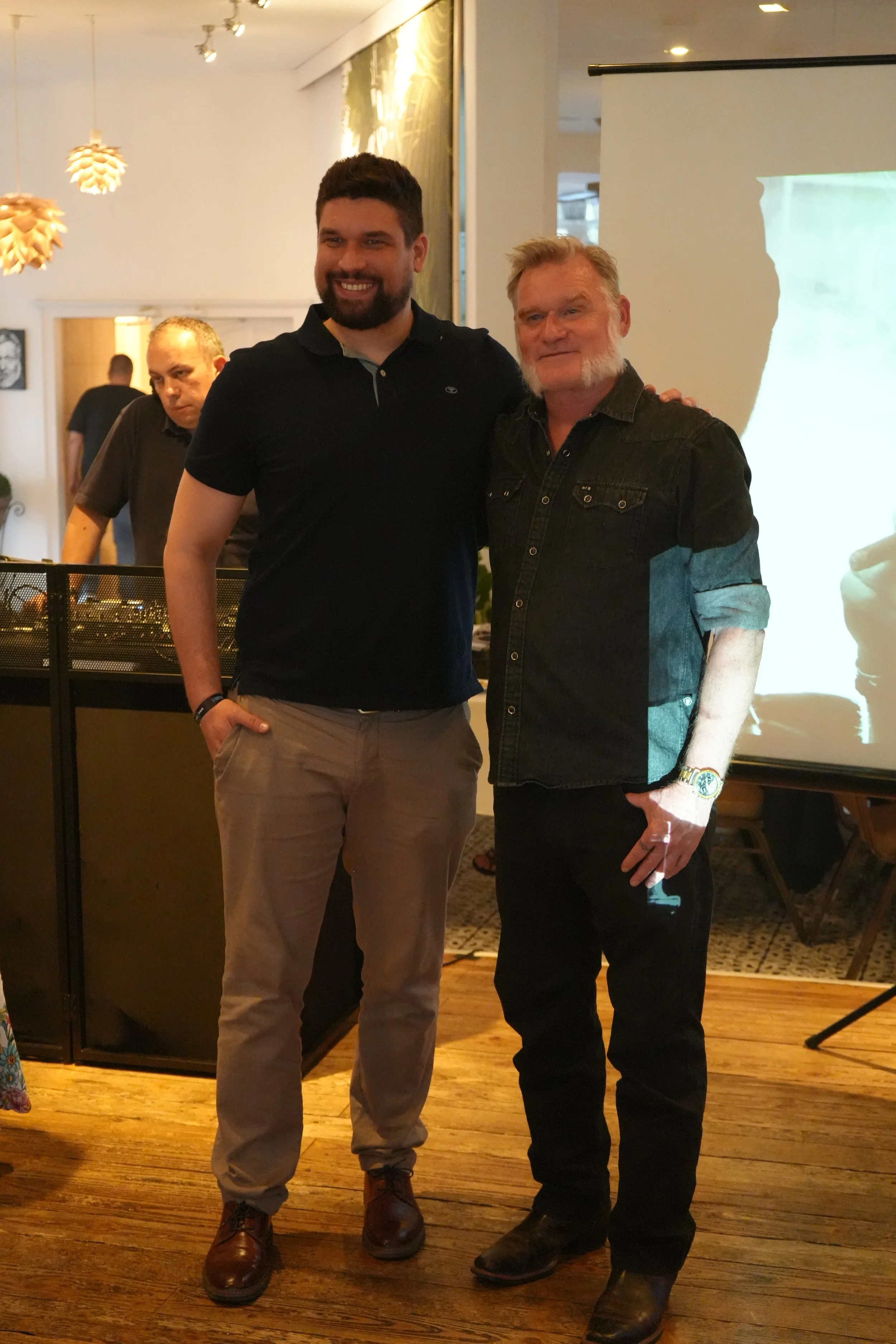 Two men standing together and smiling at an indoor event. The man on the left has dark hair and a beard, wearing a black polo shirt and khaki pants. The man on the right has light hair and a white beard, wearing a black denim shirt with rolled-up sle
