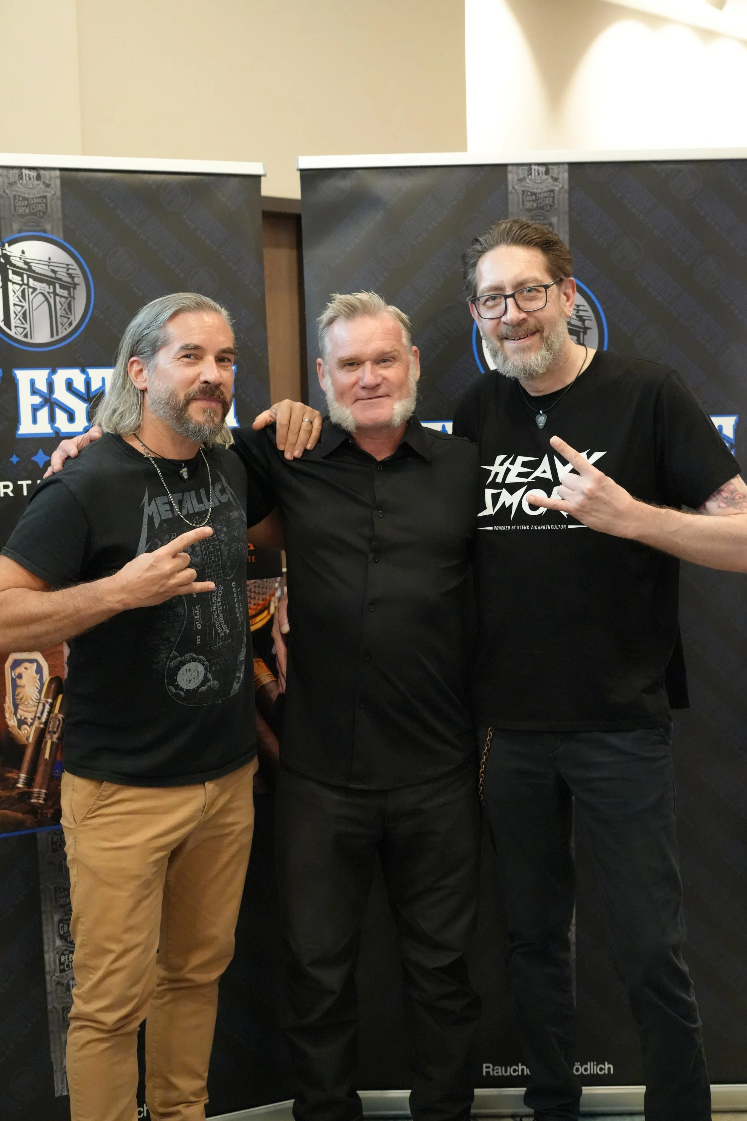 Three men with beards posing in front of black banners with blue and white logos, two wearing black t-shirts and the middle man in a black button-up shirt, all smiling and making hand gestures, at an event.