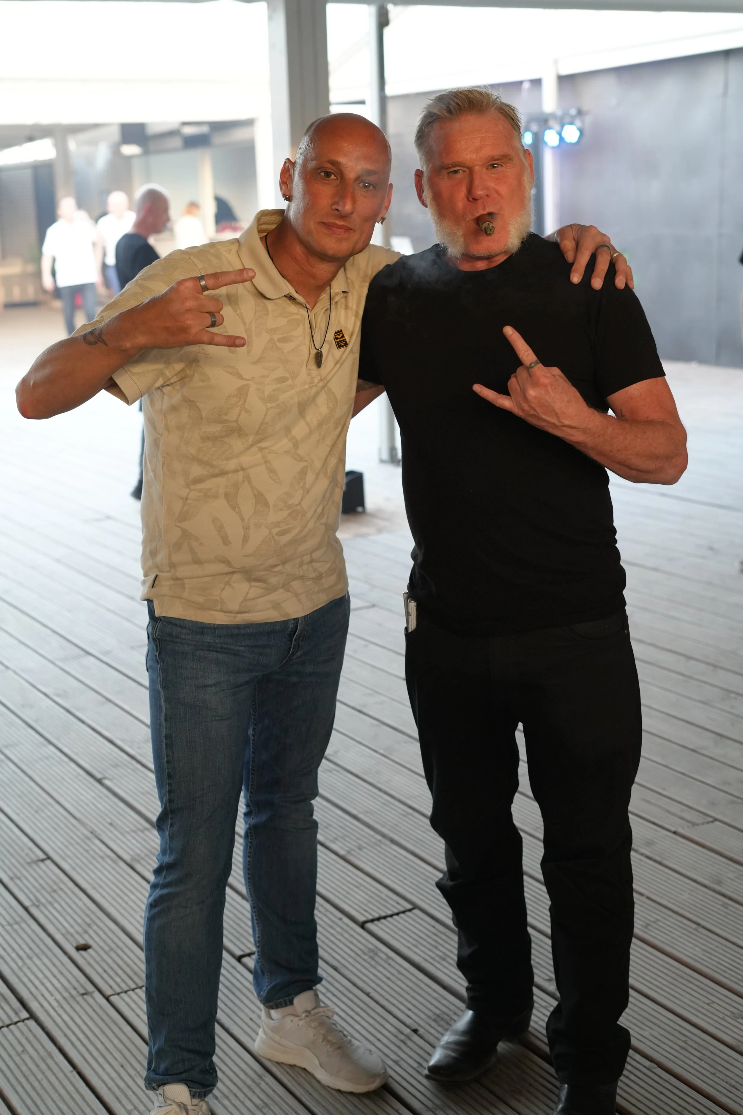 Two men standing on a wooden deck, posing for a photo at an indoor event. The man on the left is wearing a beige shirt, jeans, and white sneakers, making a rock sign with his hand. The man on the right is wearing a black shirt, black pants, and smoki