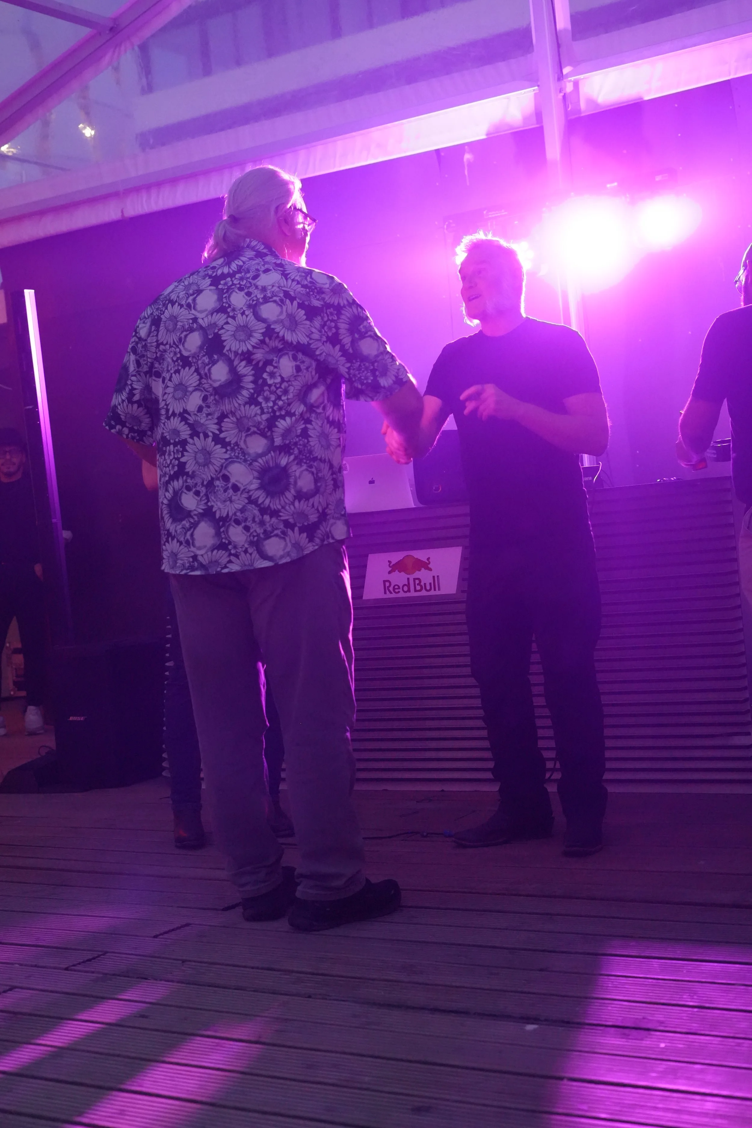 Two people shaking hands on a stage with purple lighting and a Red Bull logo in the background. One person is wearing a floral shirt, and the other is in a black shirt.